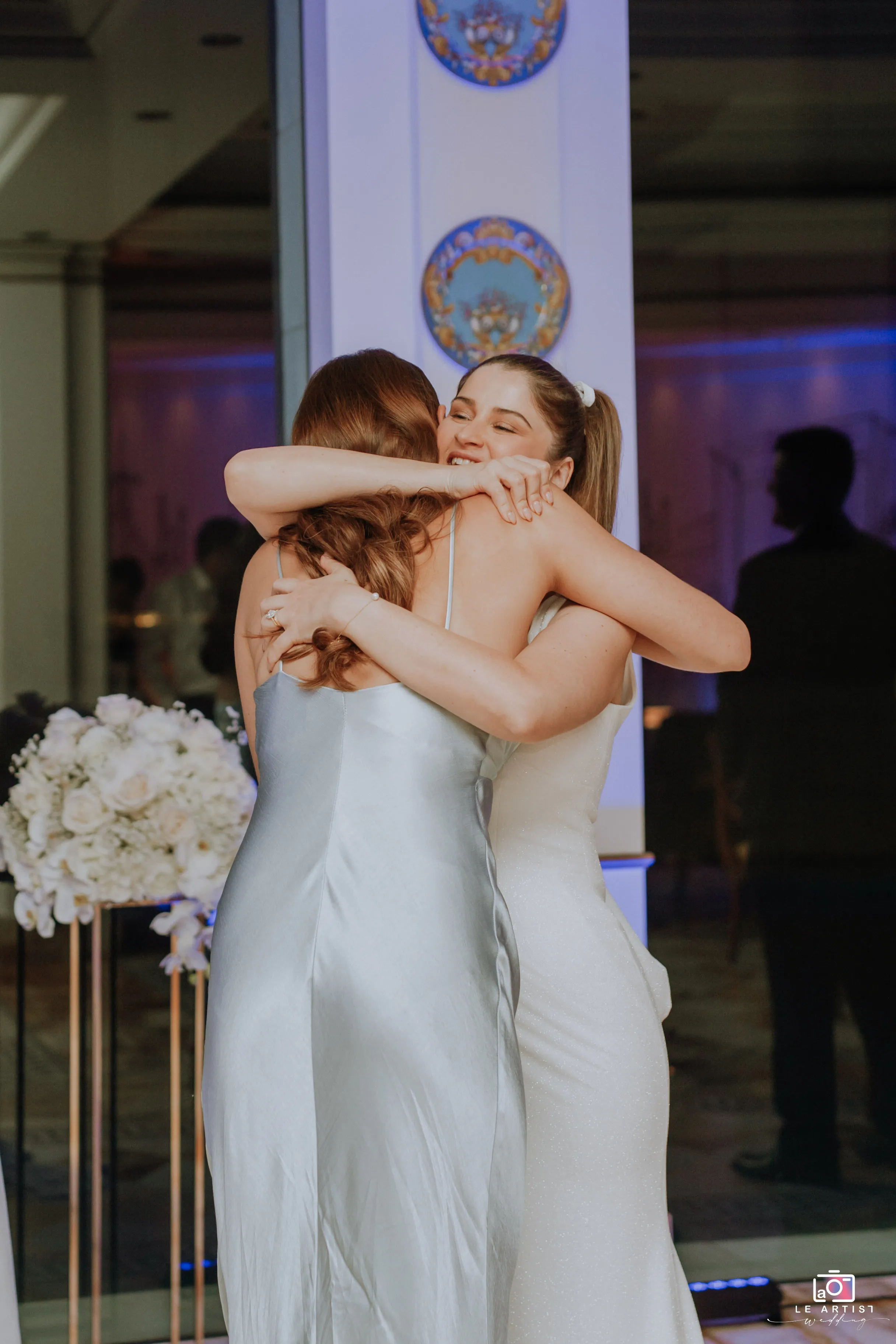A joyful embrace between the bride and a guest during the wedding reception, set against a beautifully decorated indoor backdrop. Le Artist Wedding