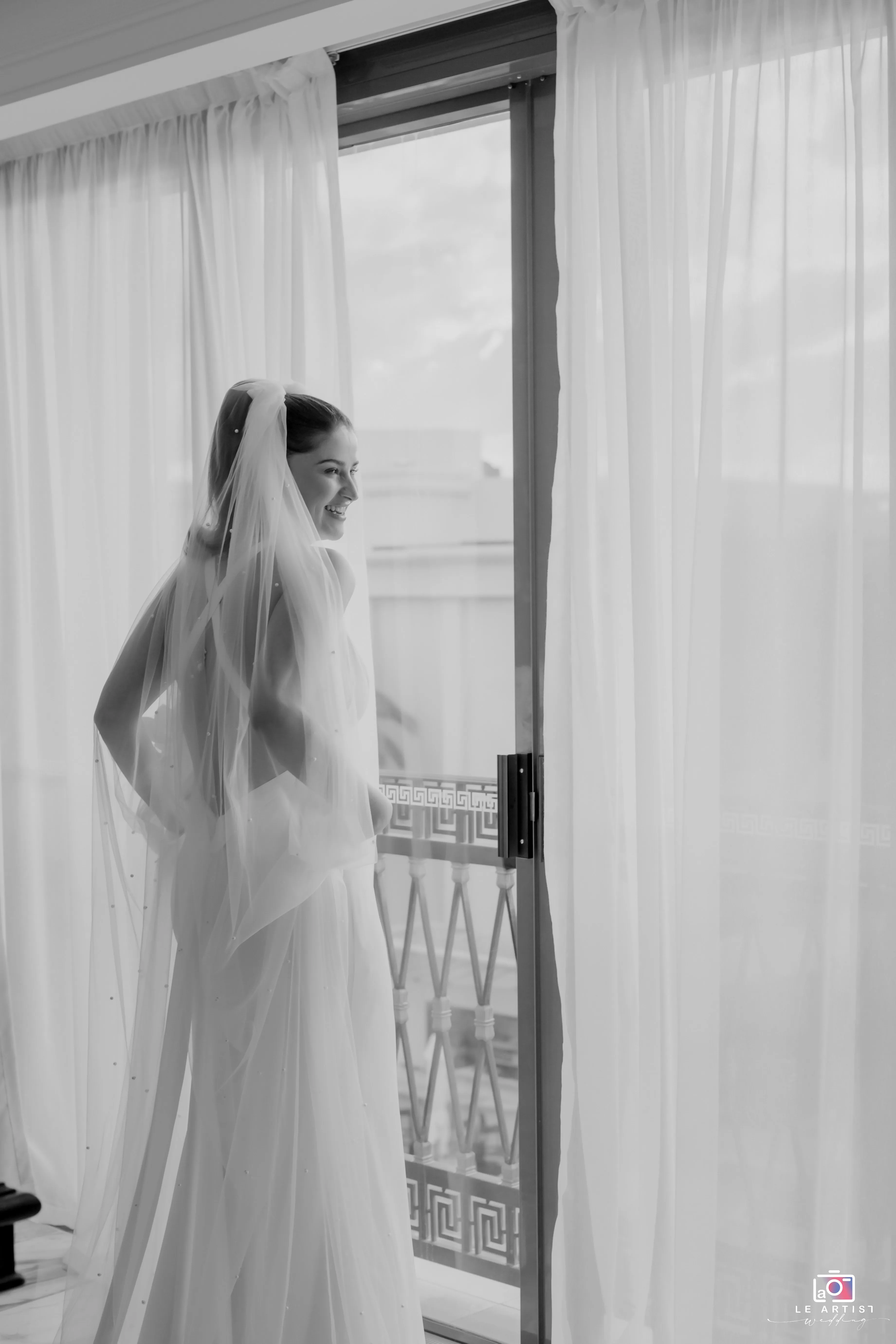 A joyful bride looks out the window during her photoshoot session, enveloped in soft, natural light. Le Artist Wedding