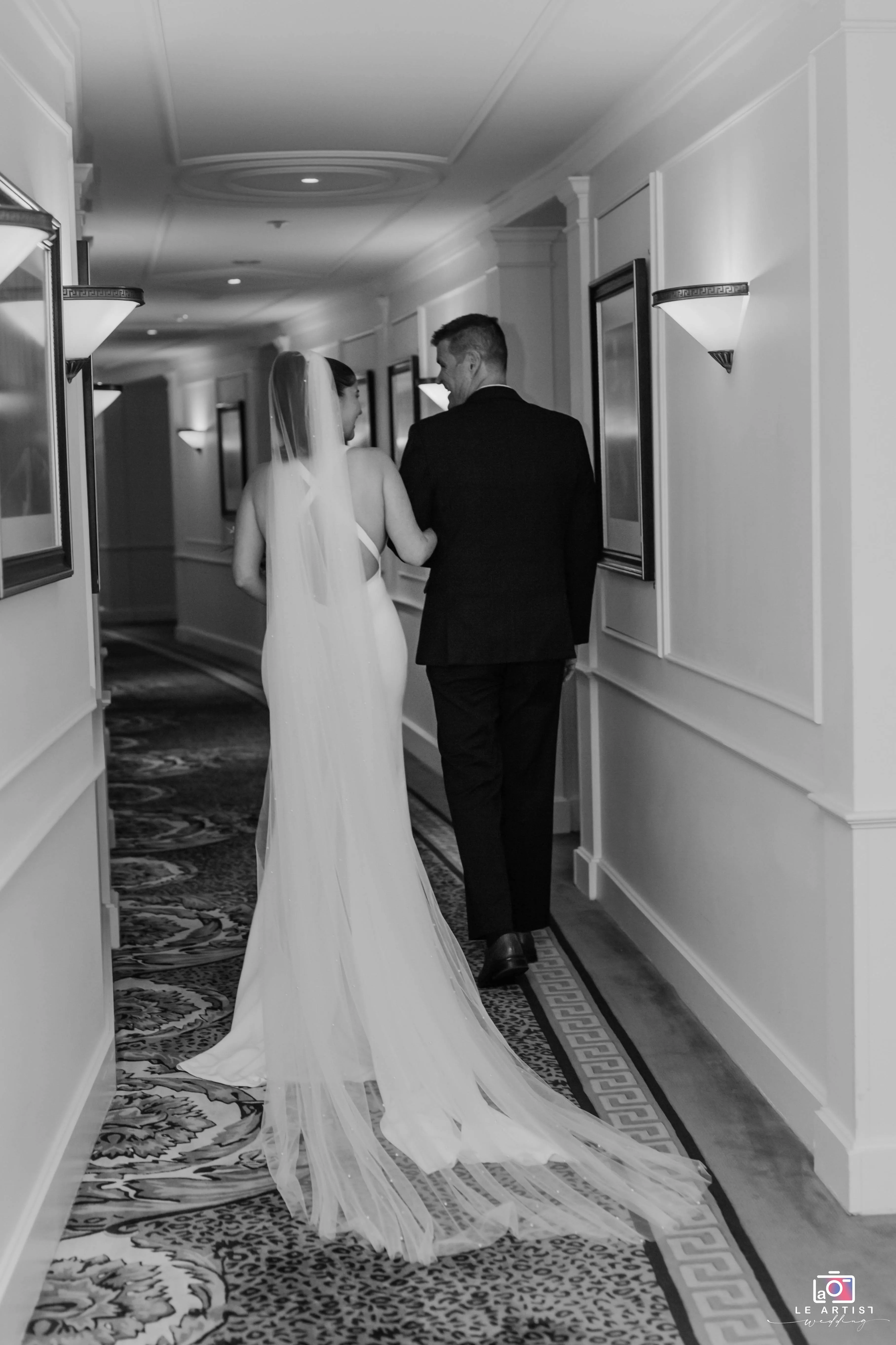 The bride walks through a hallway arm in arm with a companion, showcasing her elegant dress and veil. Le Artist Wedding