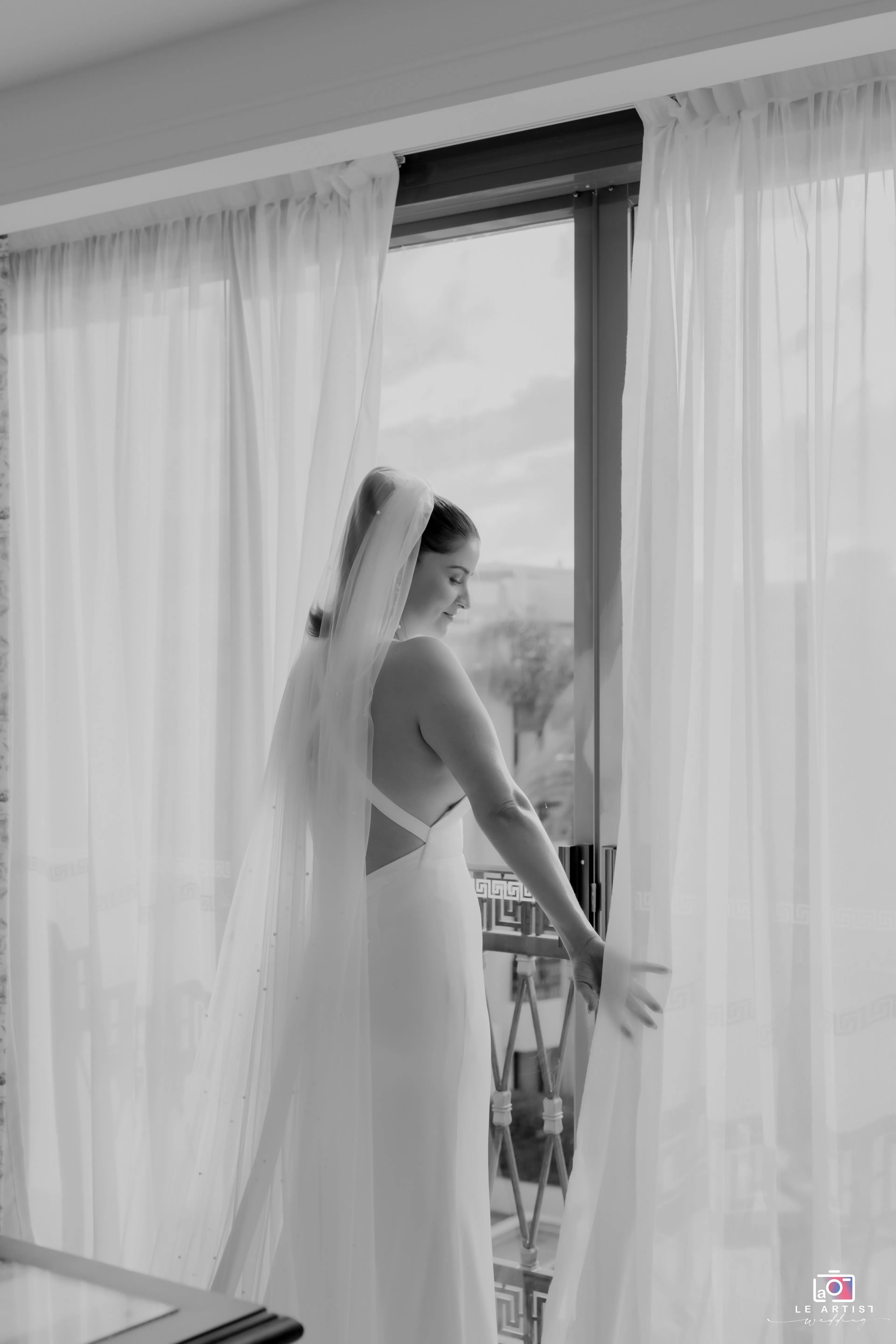 The bride stands gracefully by the window, her veil flowing behind her as she gazes outside, capturing a serene moment before the wedding ceremony. Le Artist Wedding
