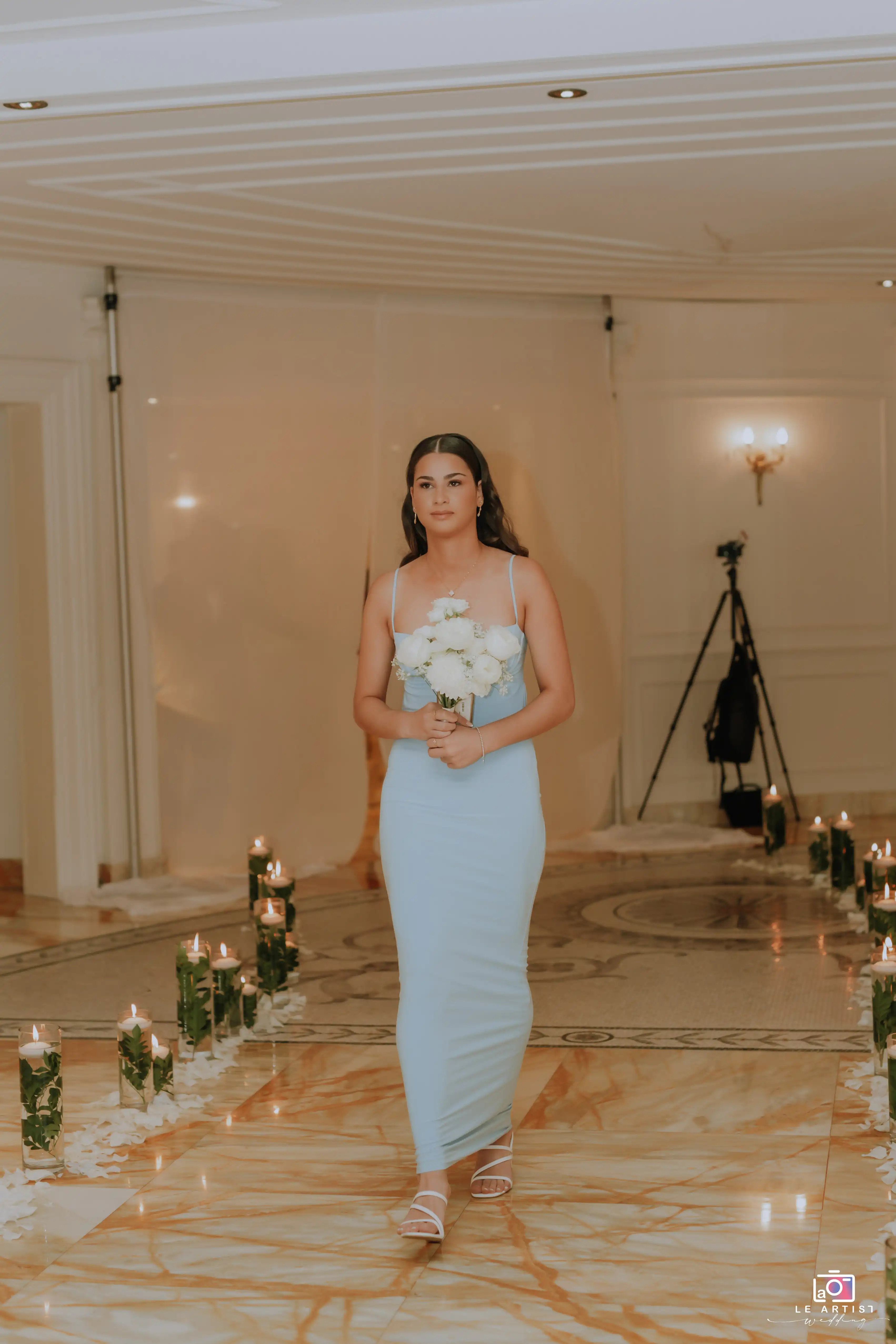 A bridesmaid walks down the aisle holding a bouquet, surrounded by candle-lit decor, during the wedding ceremony. Le Artist Wedding