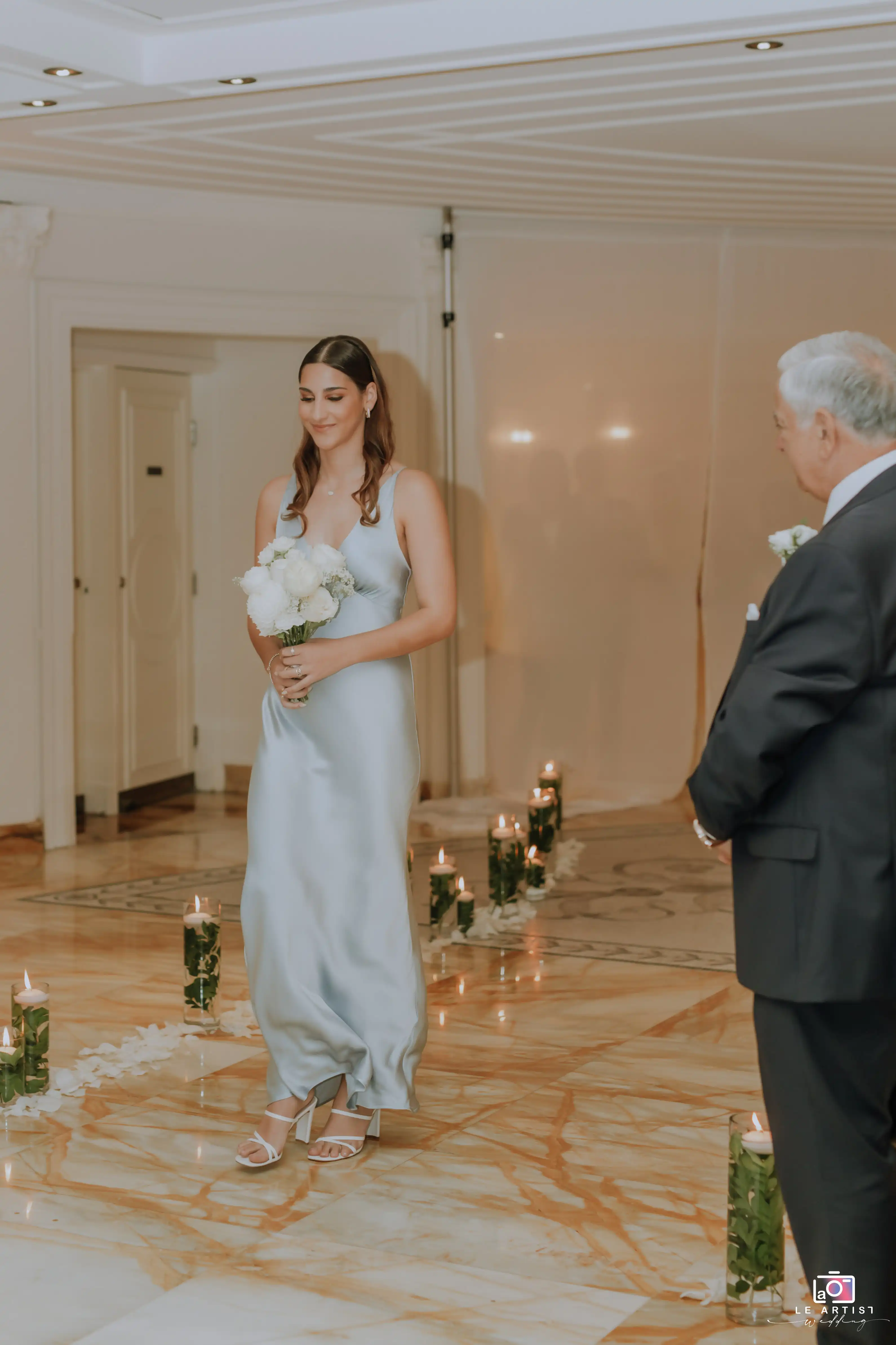 A bridesmaid walks down the aisle, carrying a bouquet during the wedding ceremony. Le Artist Wedding