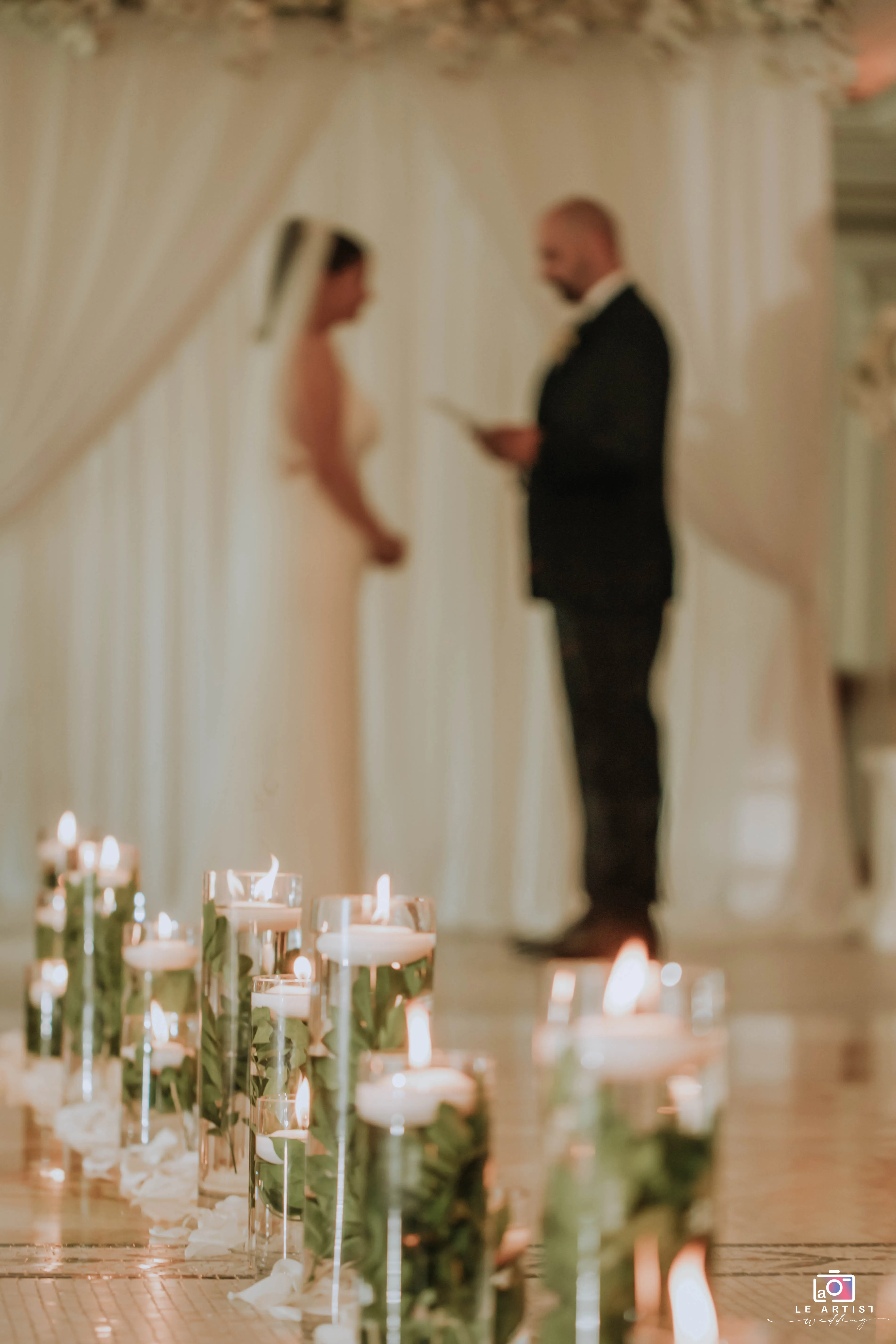 The couple stands before a backdrop of flowing drapery, exchanging vows in a candlelit setting, during their wedding ceremony. Le Artist Wedding