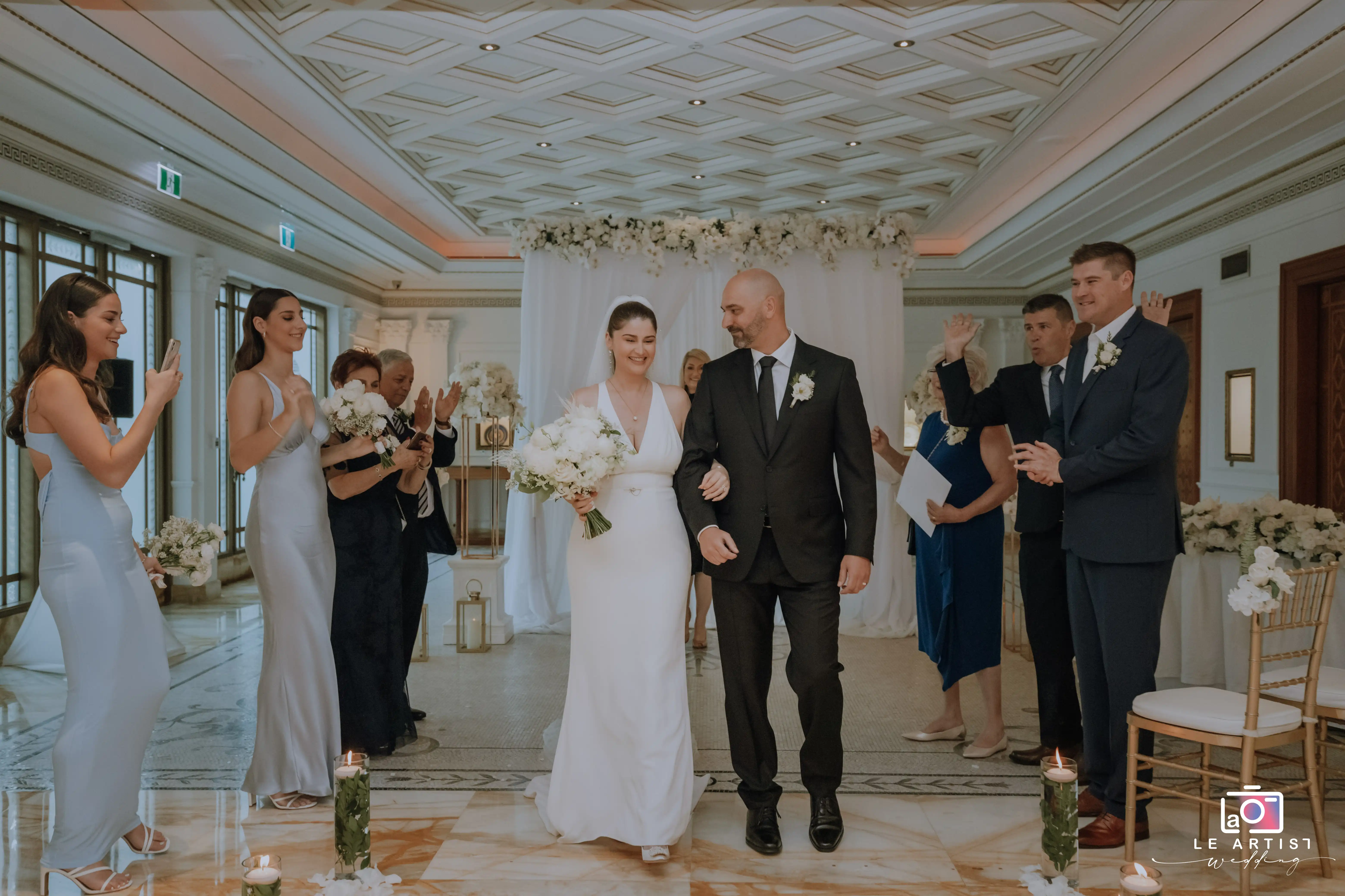 The newlyweds joyfully walk down the aisle after exchanging vows, surrounded by clapping and smiling guests, marking the conclusion of their ceremony. Le Artist Wedding