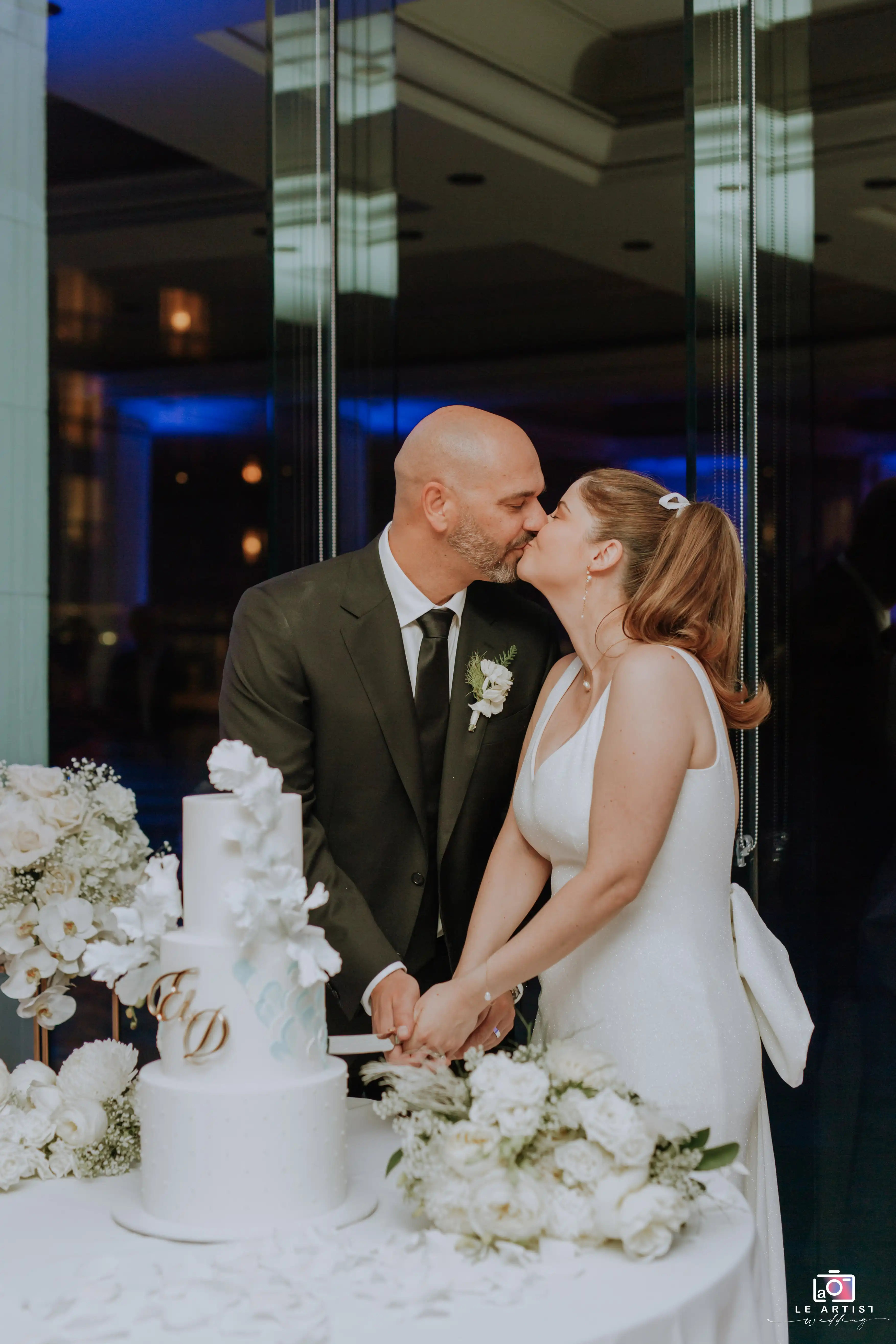 The couple shares a kiss while cutting the wedding cake during the reception celebration. Le Artist Wedding