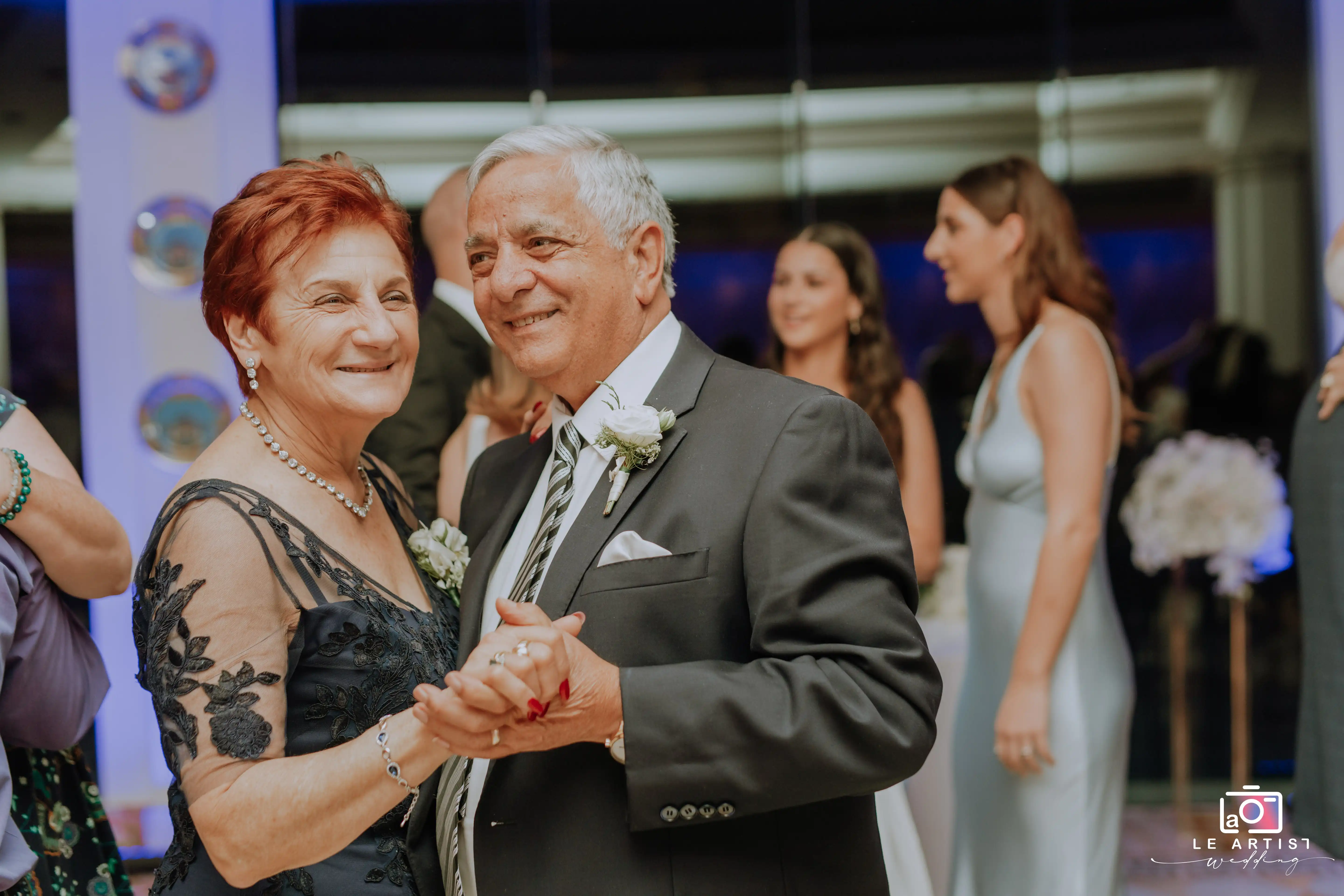 An elderly couple joyfully dances together amidst the wedding reception festivities, surrounded by smiling guests. Le Artist Wedding