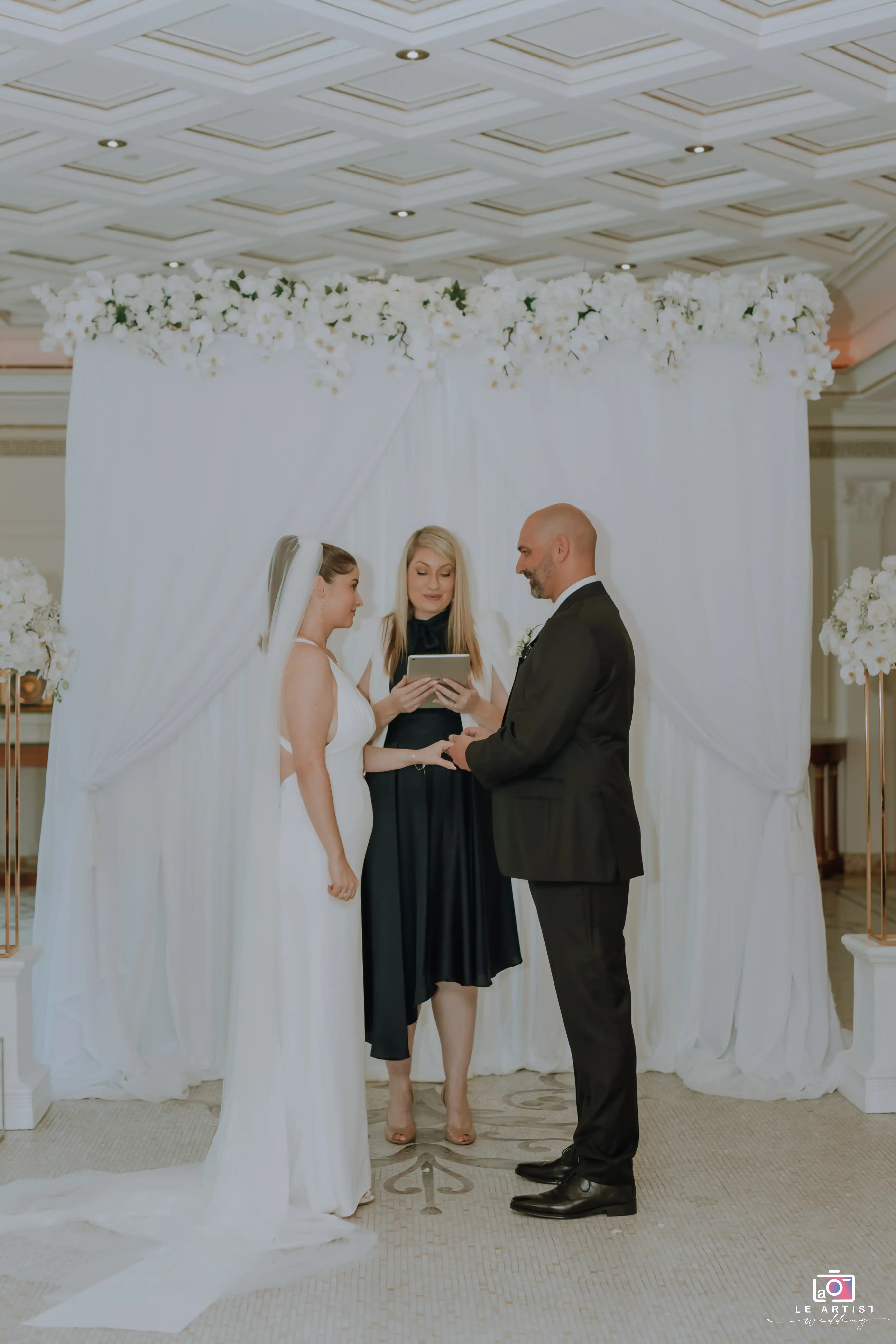 The couple exchange vows during their wedding ceremony, standing under a beautifully adorned archway. Le Artist Wedding