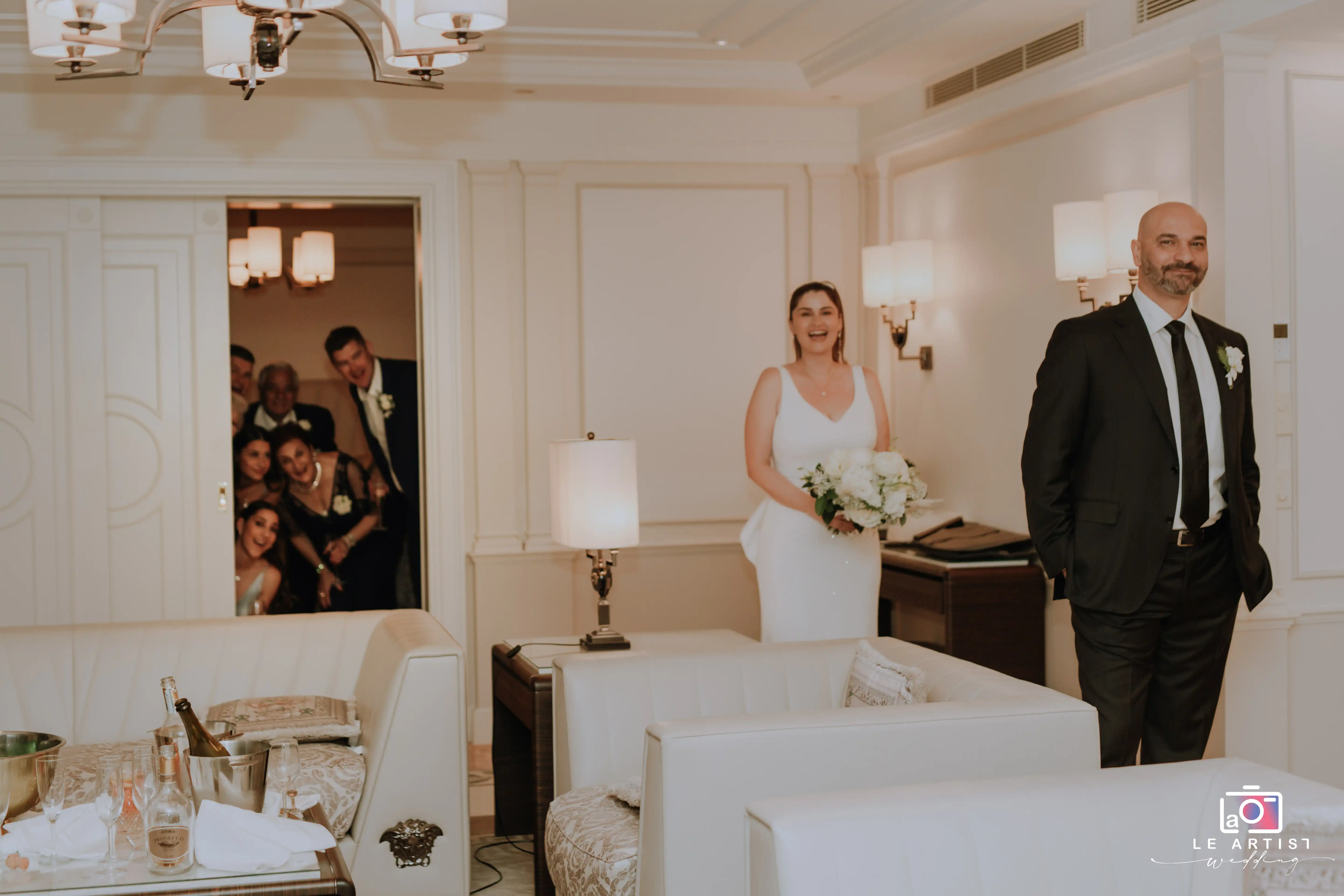 The bride and groom share a joyful moment with friends peeking in, capturing the excitement of getting ready on their big day. Le Artist Wedding