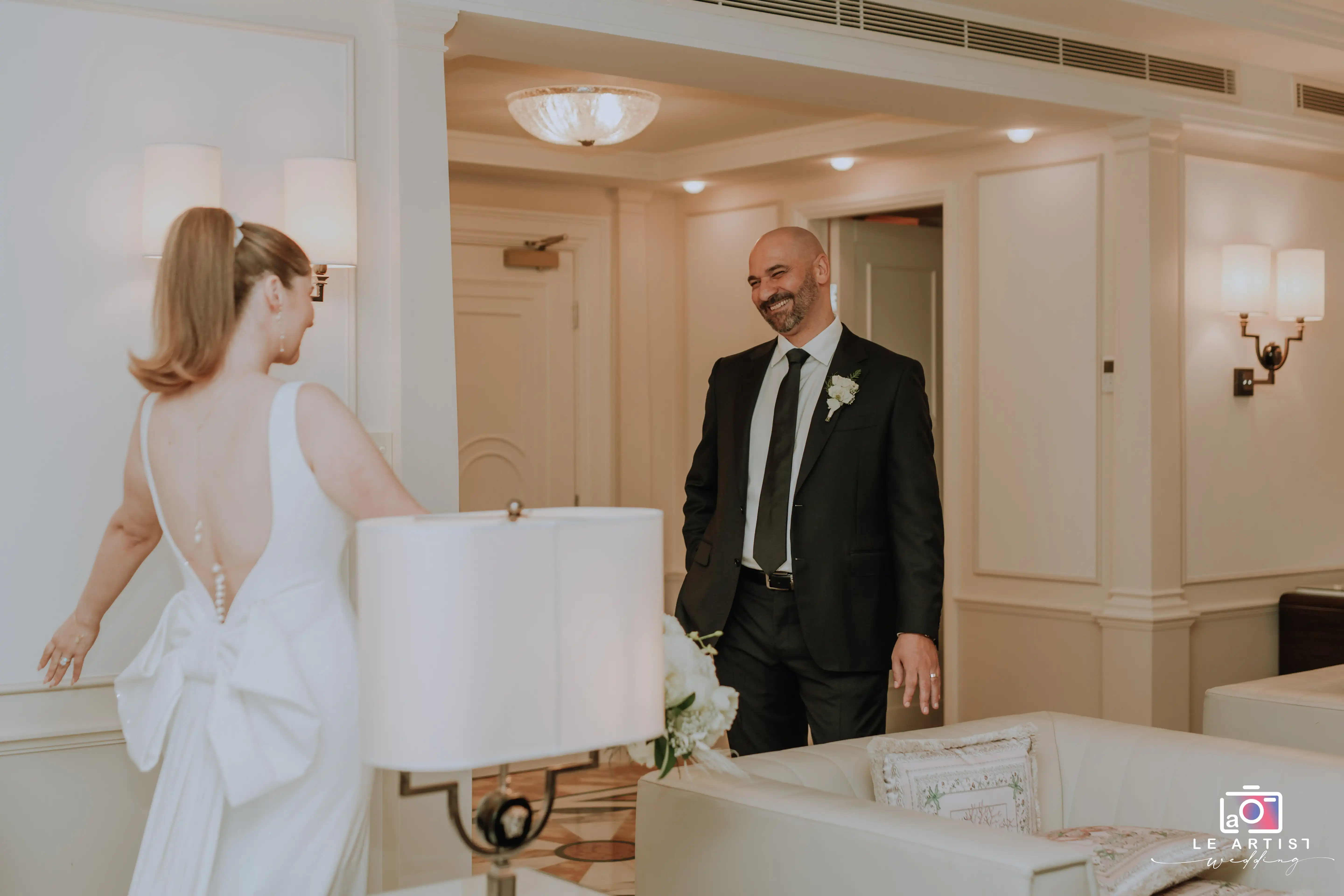 A joyful moment during the preparation phase as the bride reveals her dress to the groom, capturing the anticipation and excitement before the ceremony. Le Artist Wedding