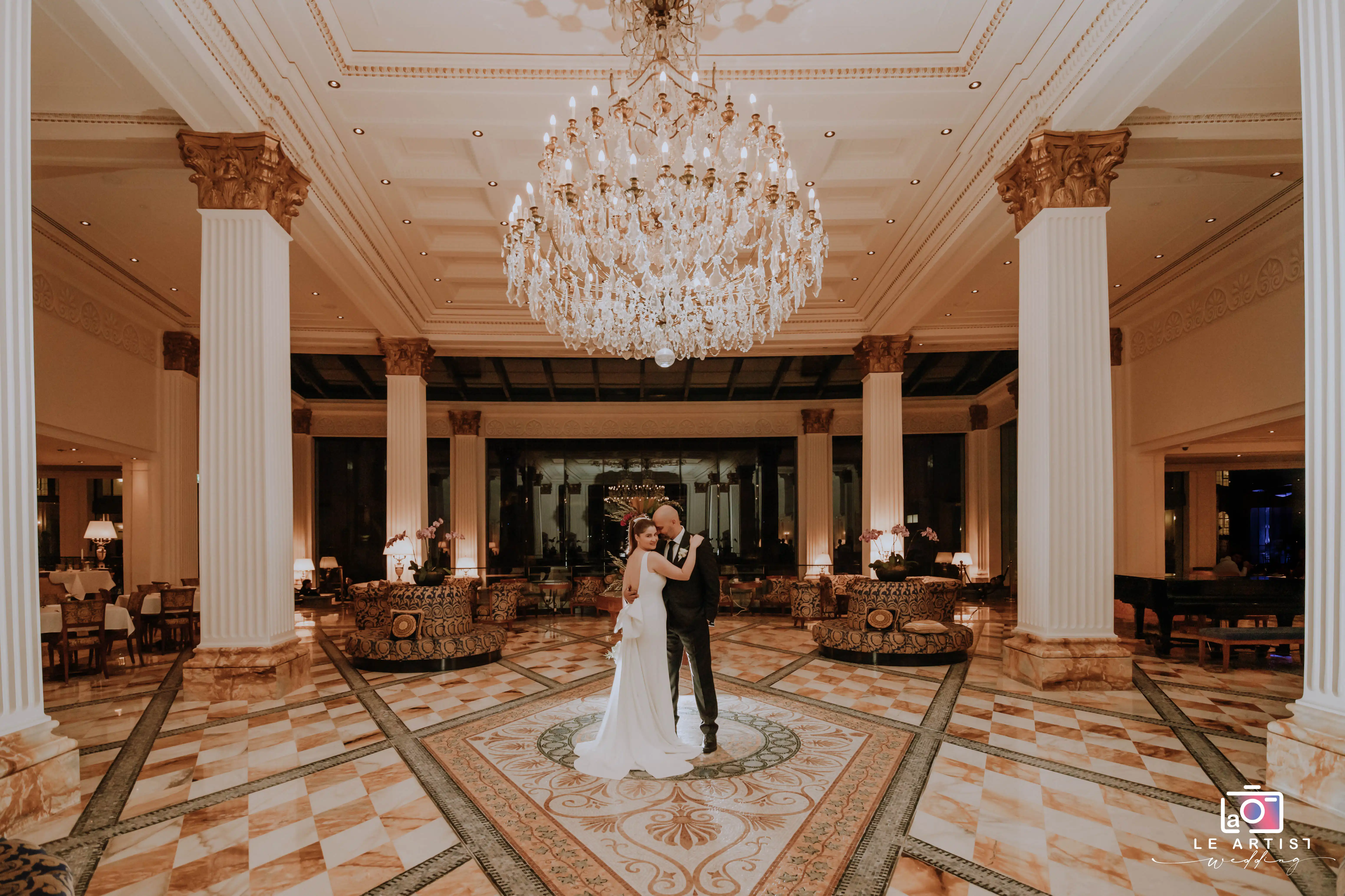 The bride and groom share a romantic moment in an opulent indoor setting under a grand chandelier. Le Artist Wedding