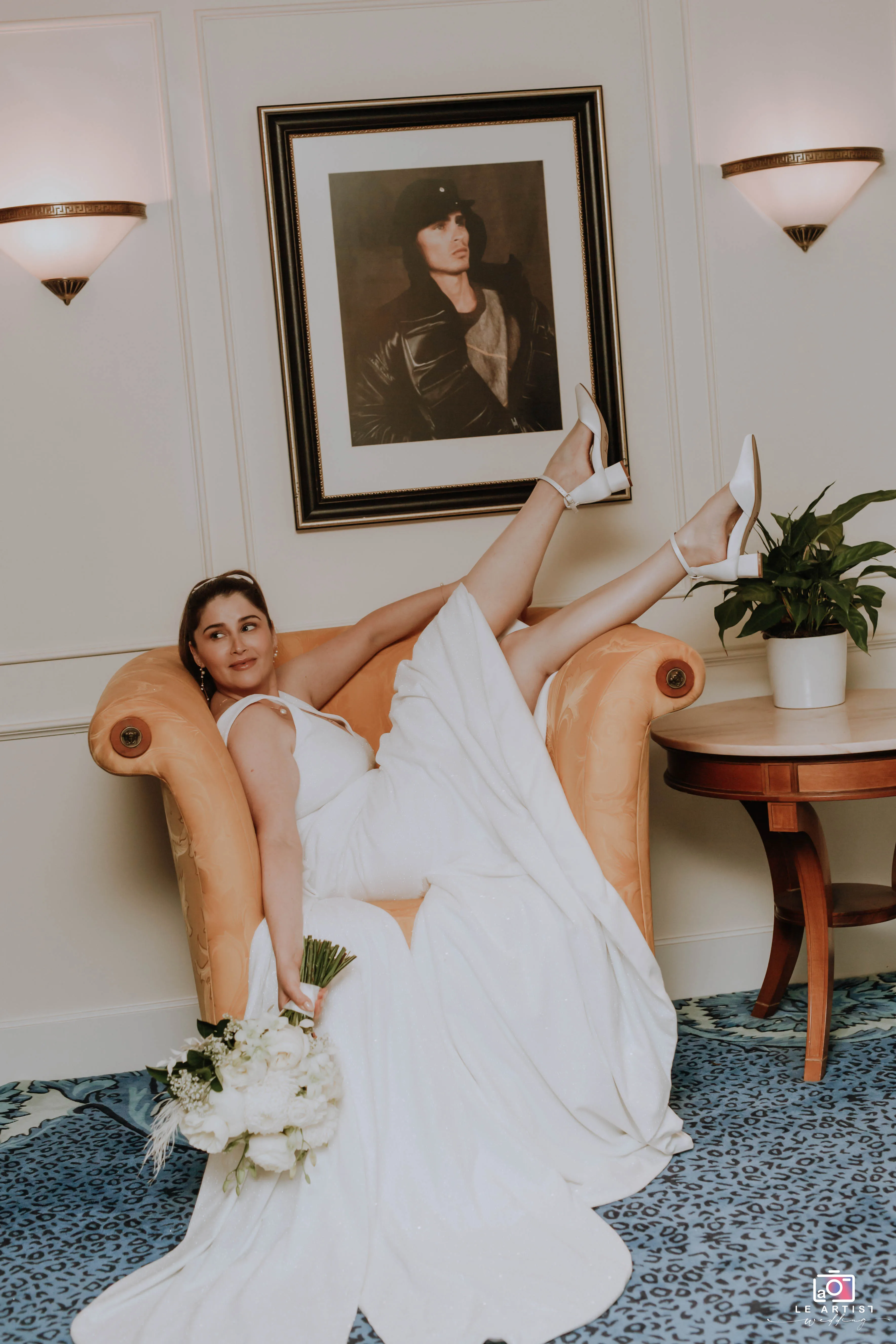 The bride strikes a playful pose on a luxurious armchair, showcasing her elegant dress and bouquet. Le Artist Wedding