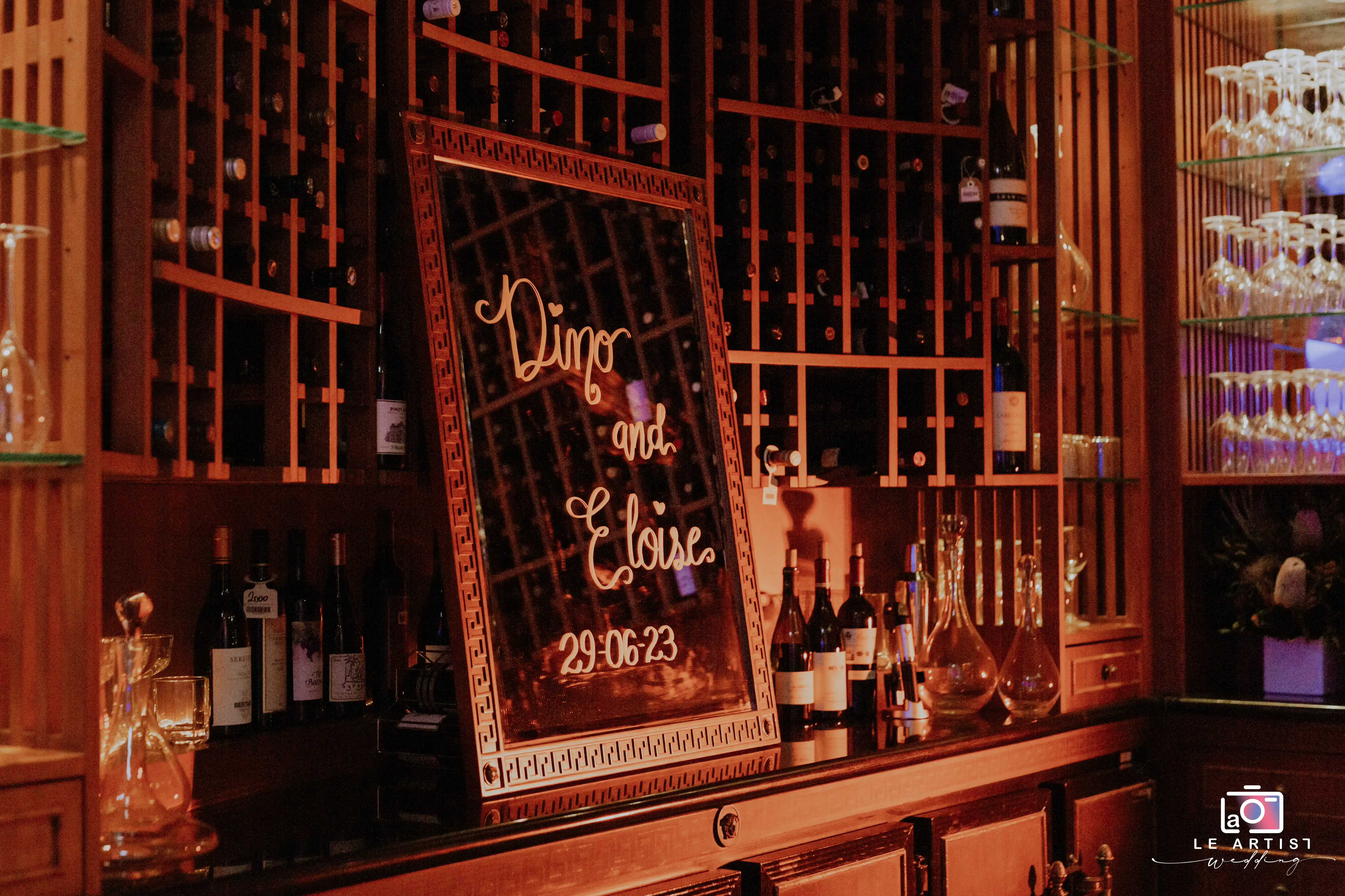 A stylish display at the wedding reception bar area, featuring a personalized mirror sign with the couple's names and wedding date. Le Artist Wedding