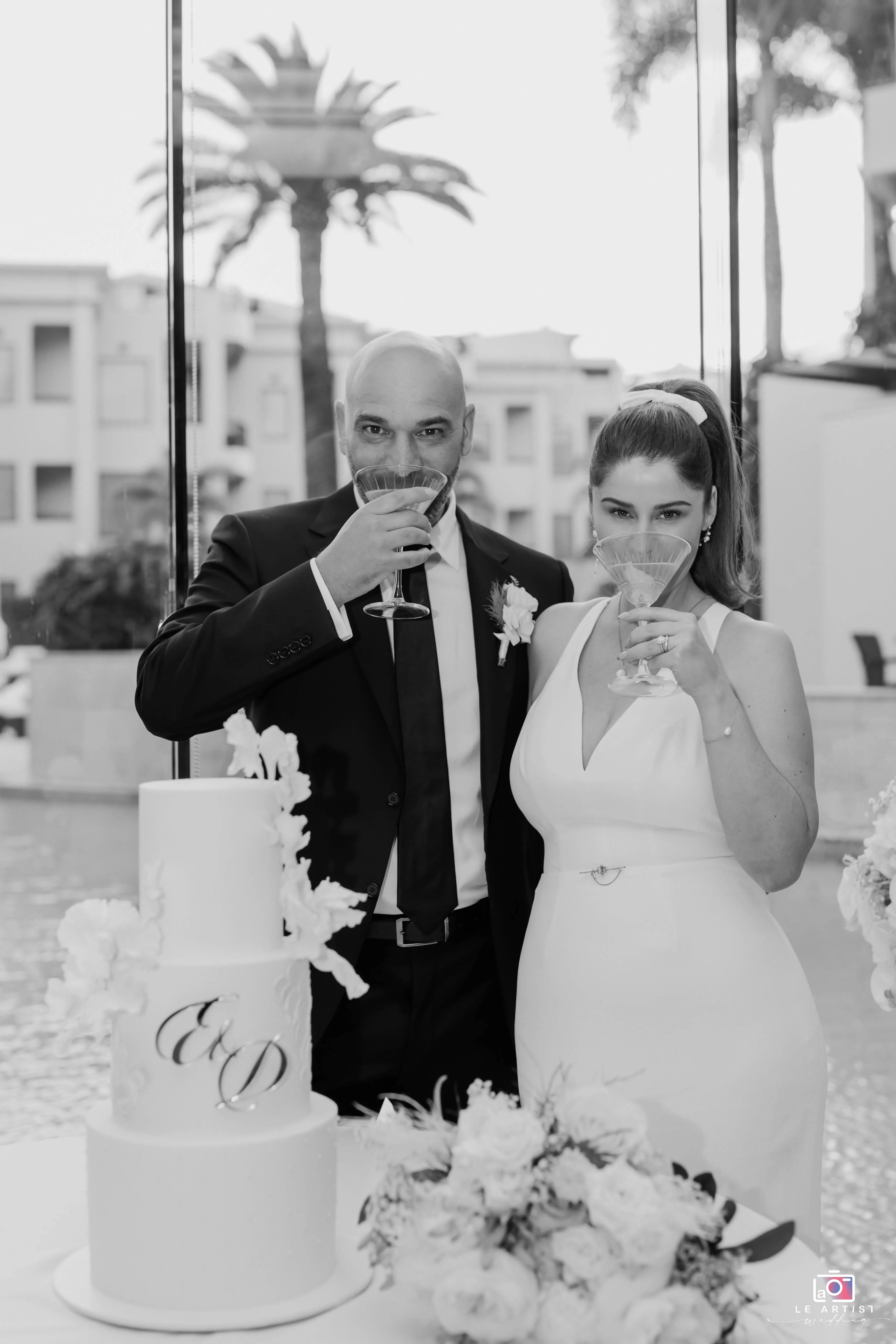 The newlyweds share a toast by their elegantly decorated wedding cake, celebrating their union at the reception. Le Artist Wedding