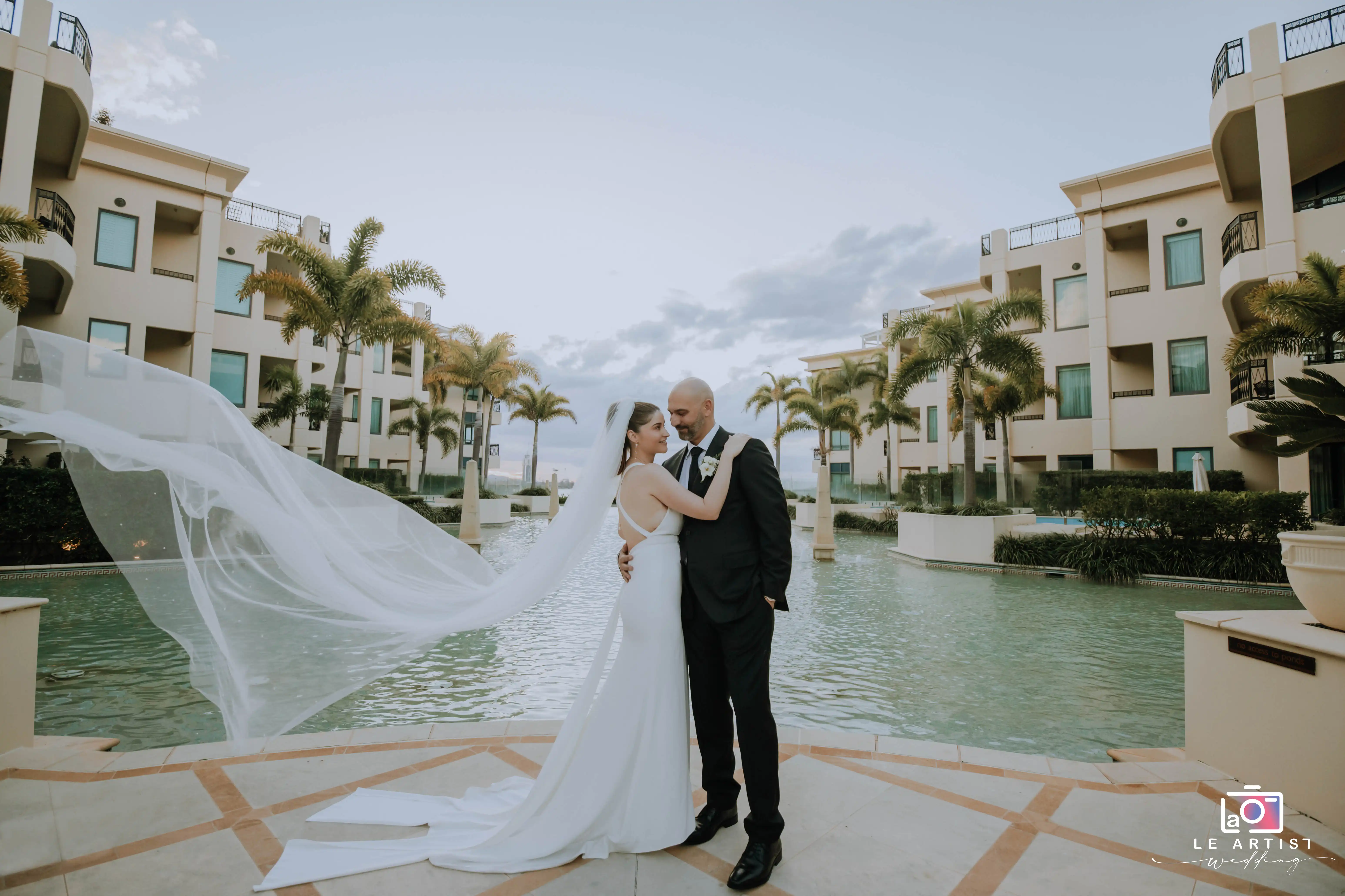 The bride and groom embrace by a serene pool, with the bride's veil softly flowing in the breeze, capturing a romantic moment against the backdrop of architectural elegance and palm trees. Le Artist Wedding
