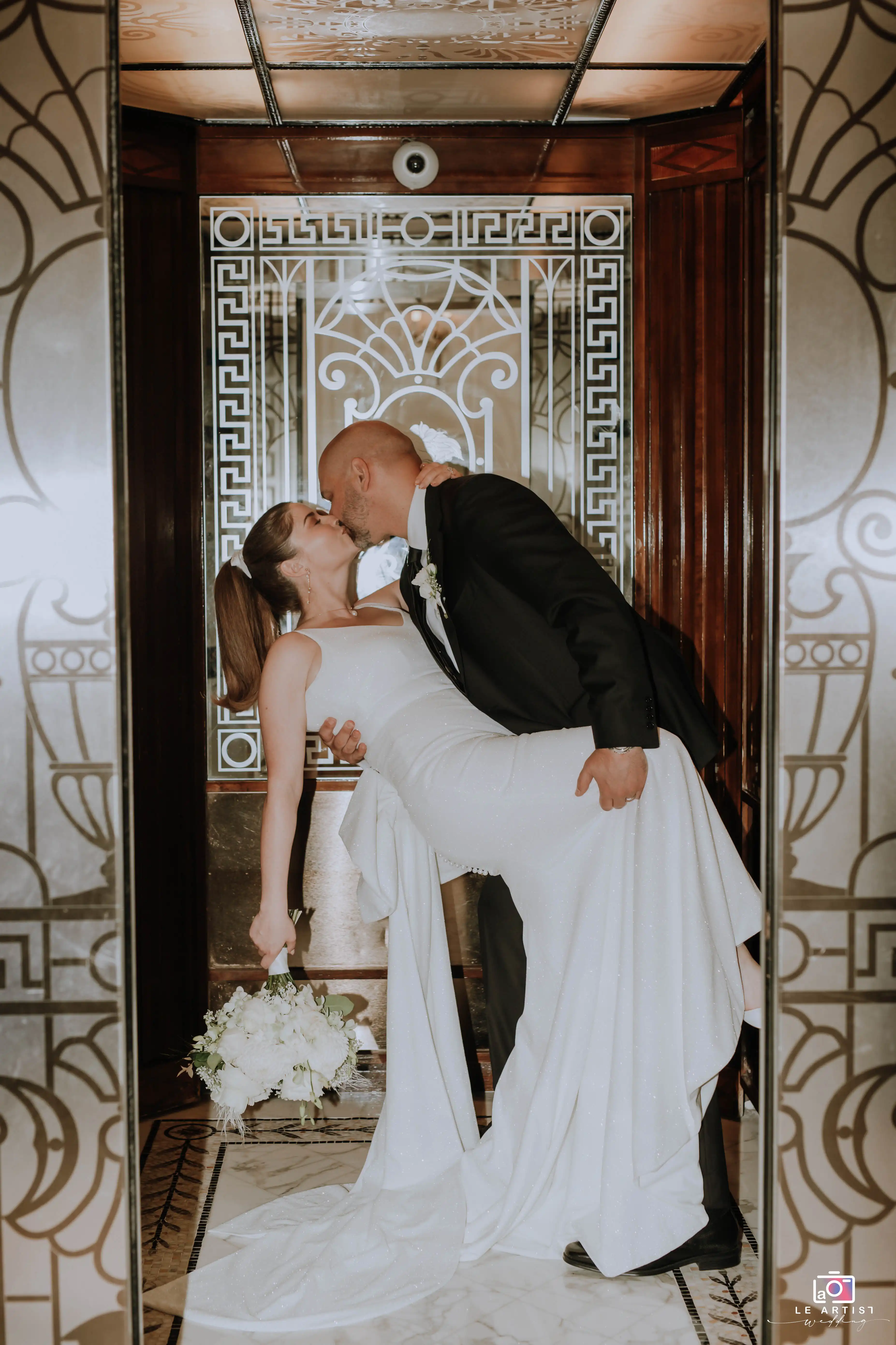 A romantic moment captured during the bridal photoshoot as the groom holds the bride in an elegant pose inside an ornately decorated elevator. Le Artist Wedding
