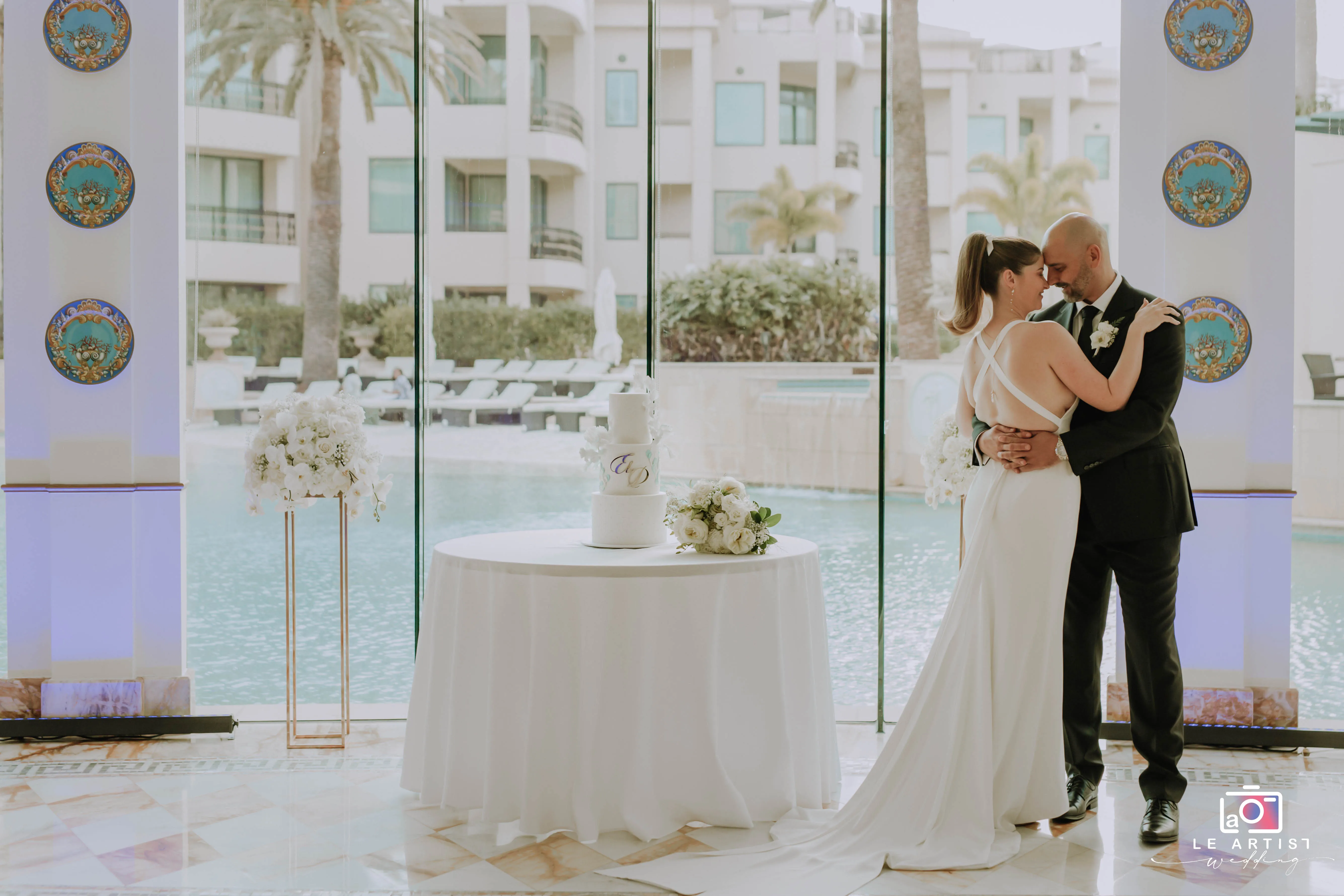 A romantic moment between the bride and groom captured indoors, with a stunning view of the poolside, framed by elegant decor and a beautifully set cake table. Le Artist Wedding