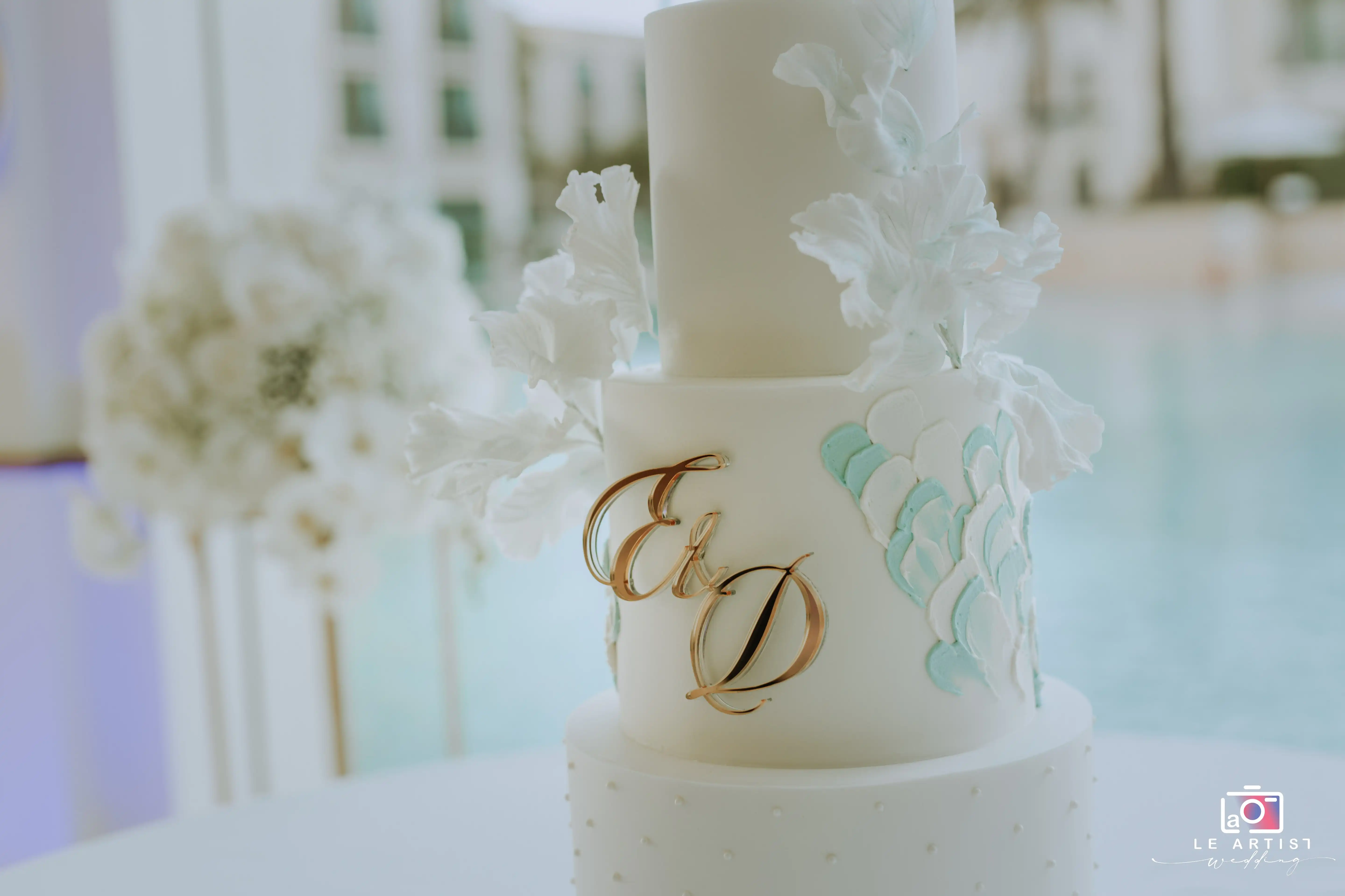 A beautifully designed wedding cake adorned with elegant white flowers and initials, perfectly staged for the celebration at the reception. Le Artist Wedding