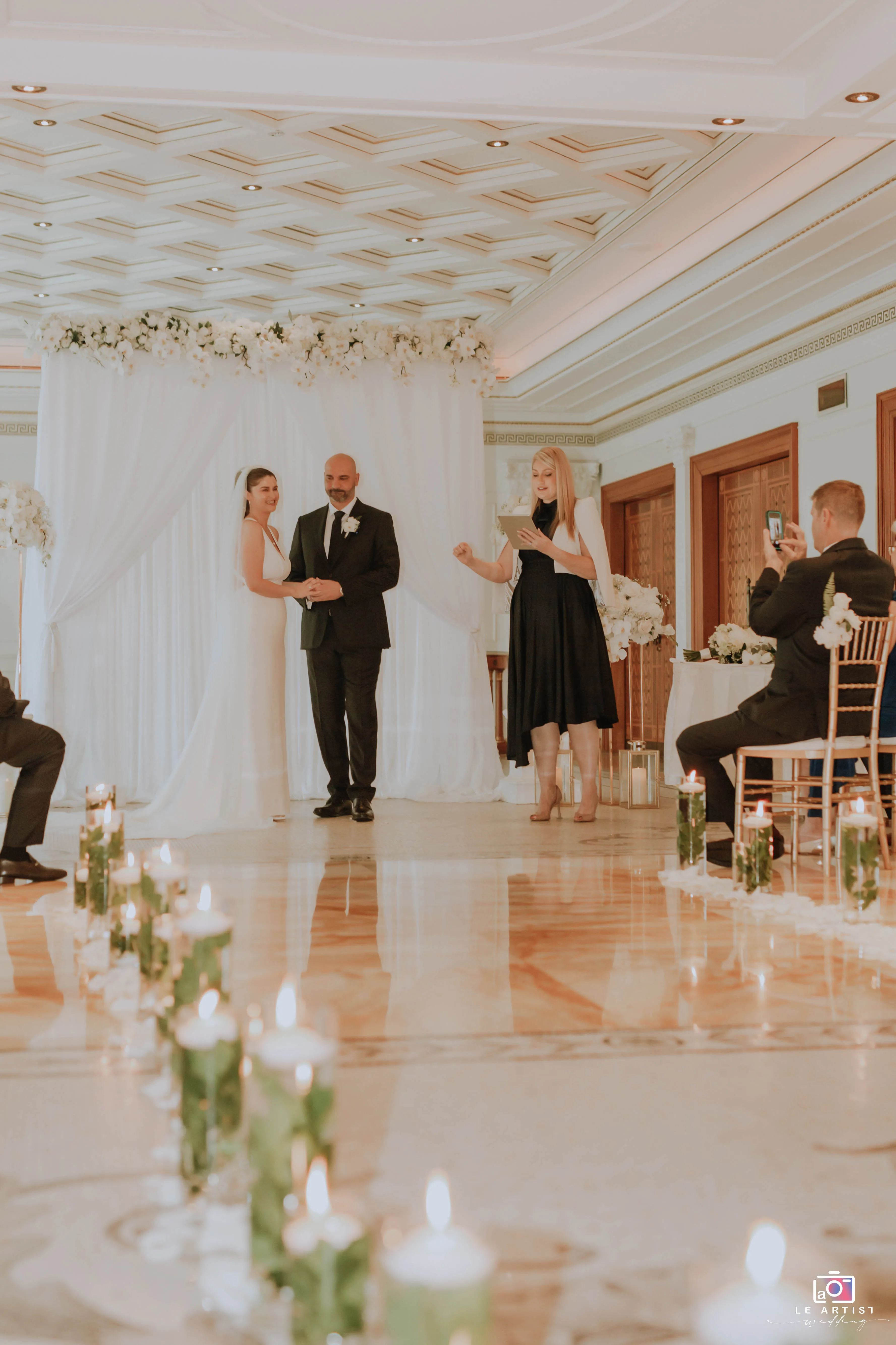 The couple exchanges vows in an elegantly decorated venue, surrounded by guests and soft candlelight during their wedding ceremony. Le Artist Wedding