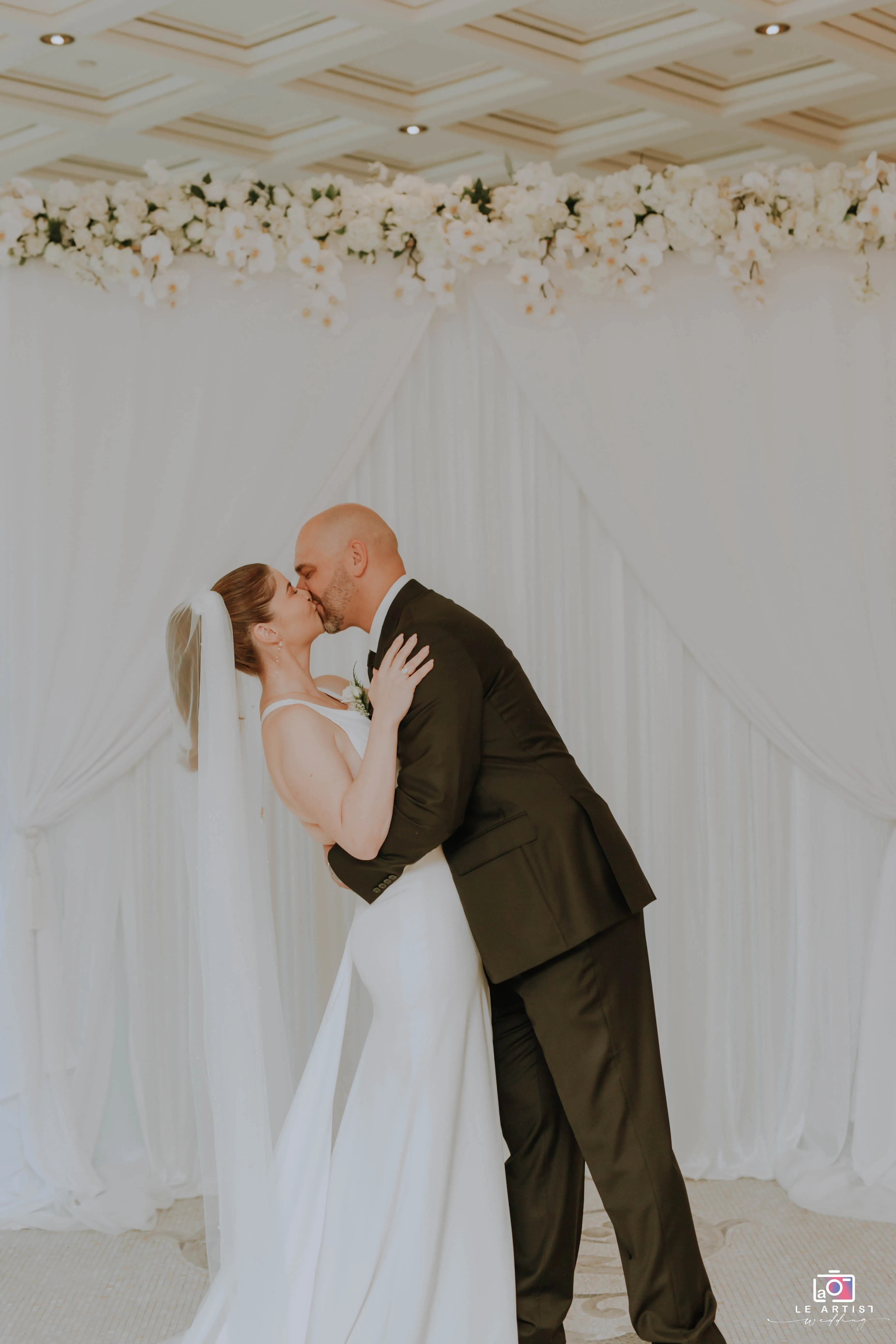 The couple shares a joyful kiss during their wedding ceremony under a beautifully decorated arch. Le Artist Wedding