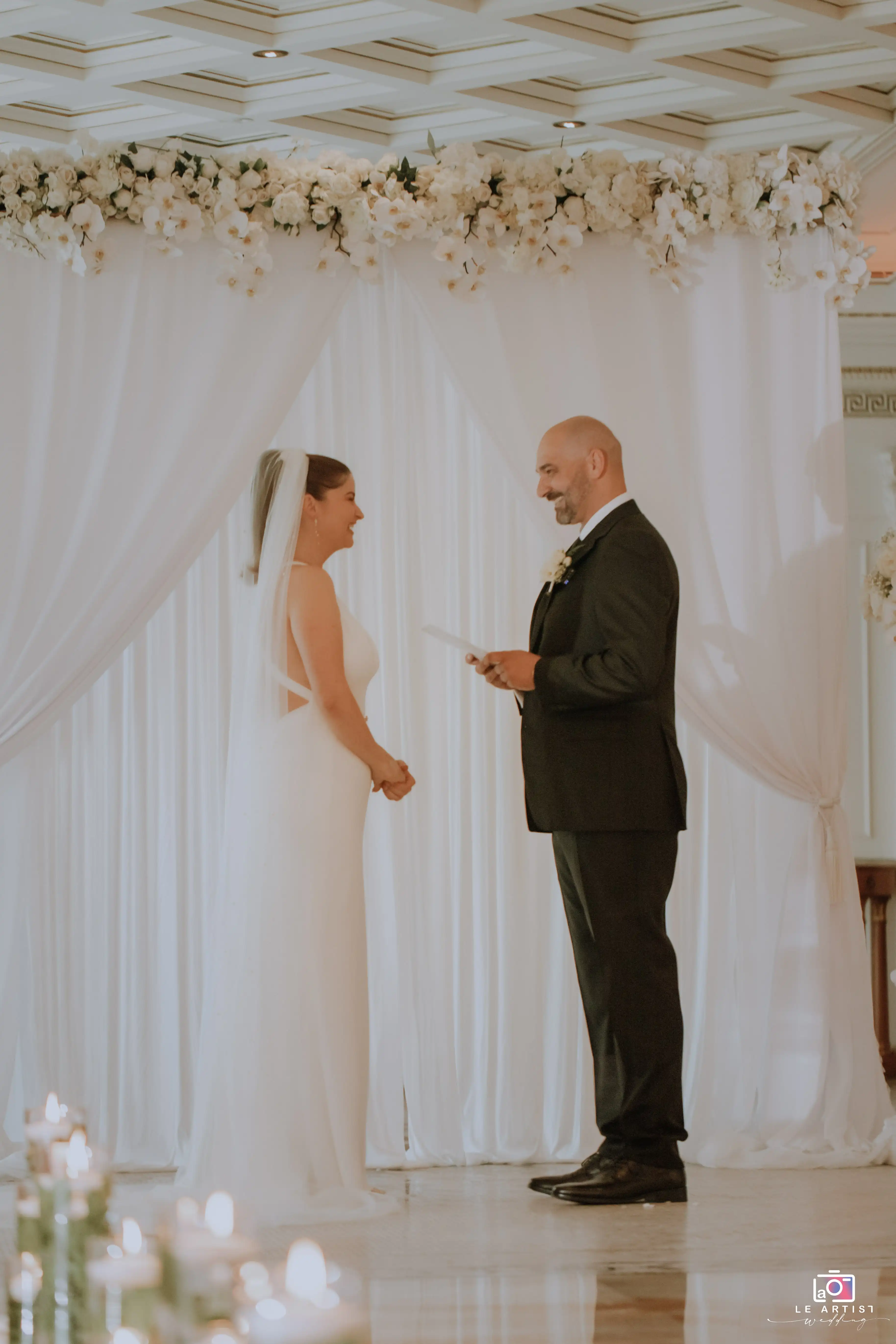 The bride and groom exchange heartfelt vows surrounded by elegant floral arrangements during their wedding ceremony. Le Artist Wedding