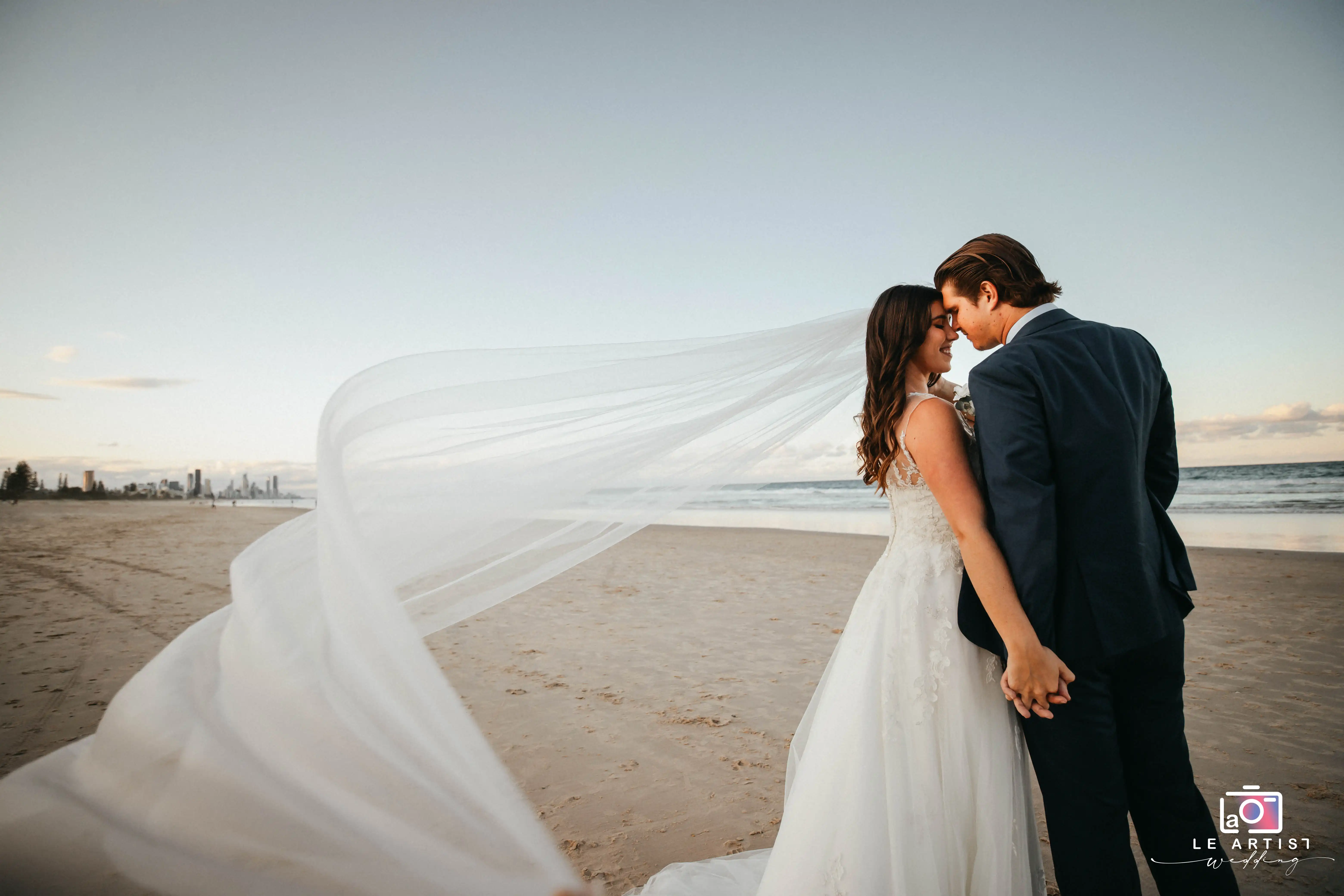 A romantic moment captured as the couple holds hands, embracing on a serene beach. The bride's veil flows gracefully in the breeze, adding to the picturesque scene with the ocean and skyline in the background. Le Artist Wedding