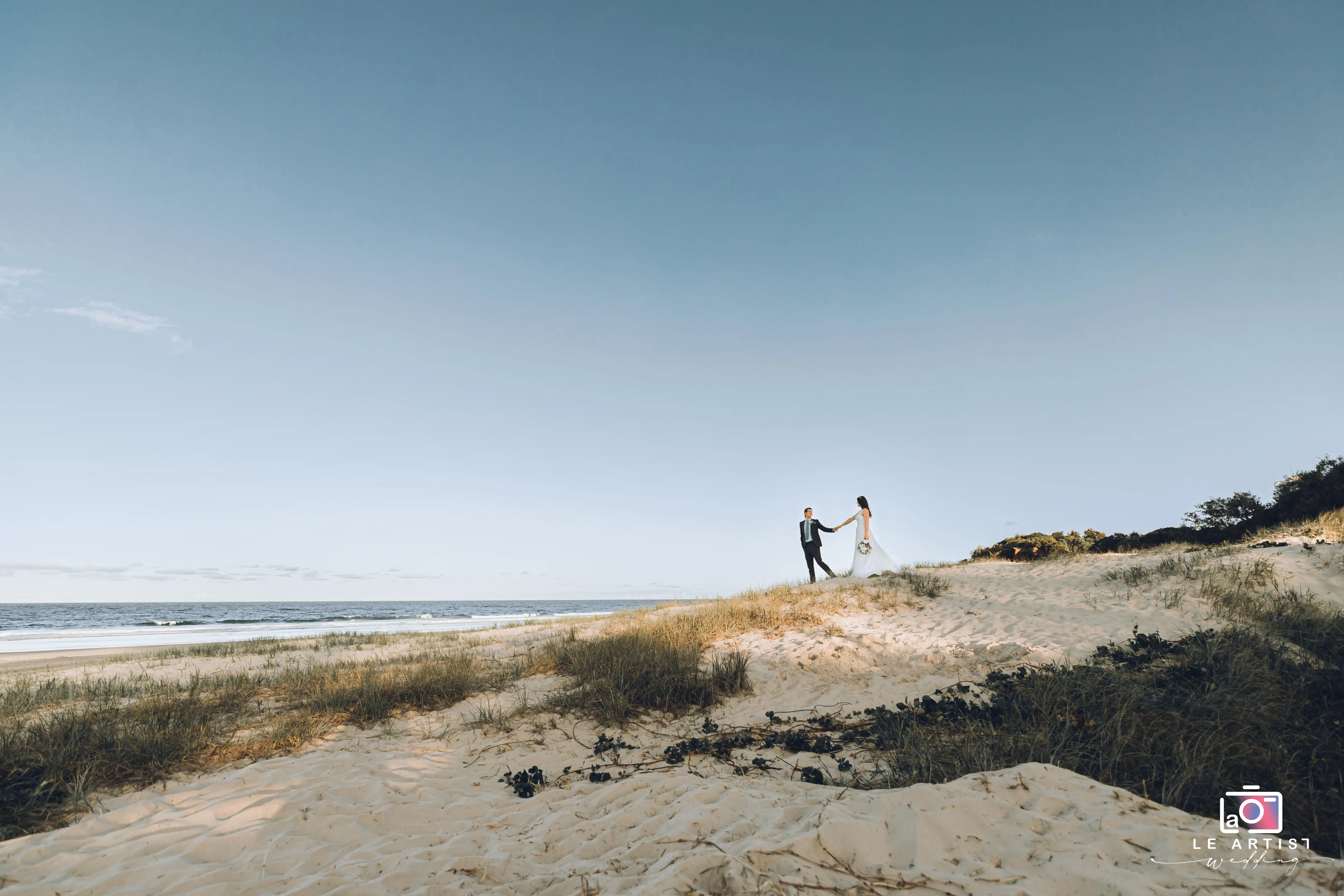 A couple stands hand in hand on a serene beach dune, set against a vast sky, symbolizing their journey together. Le Artist Wedding