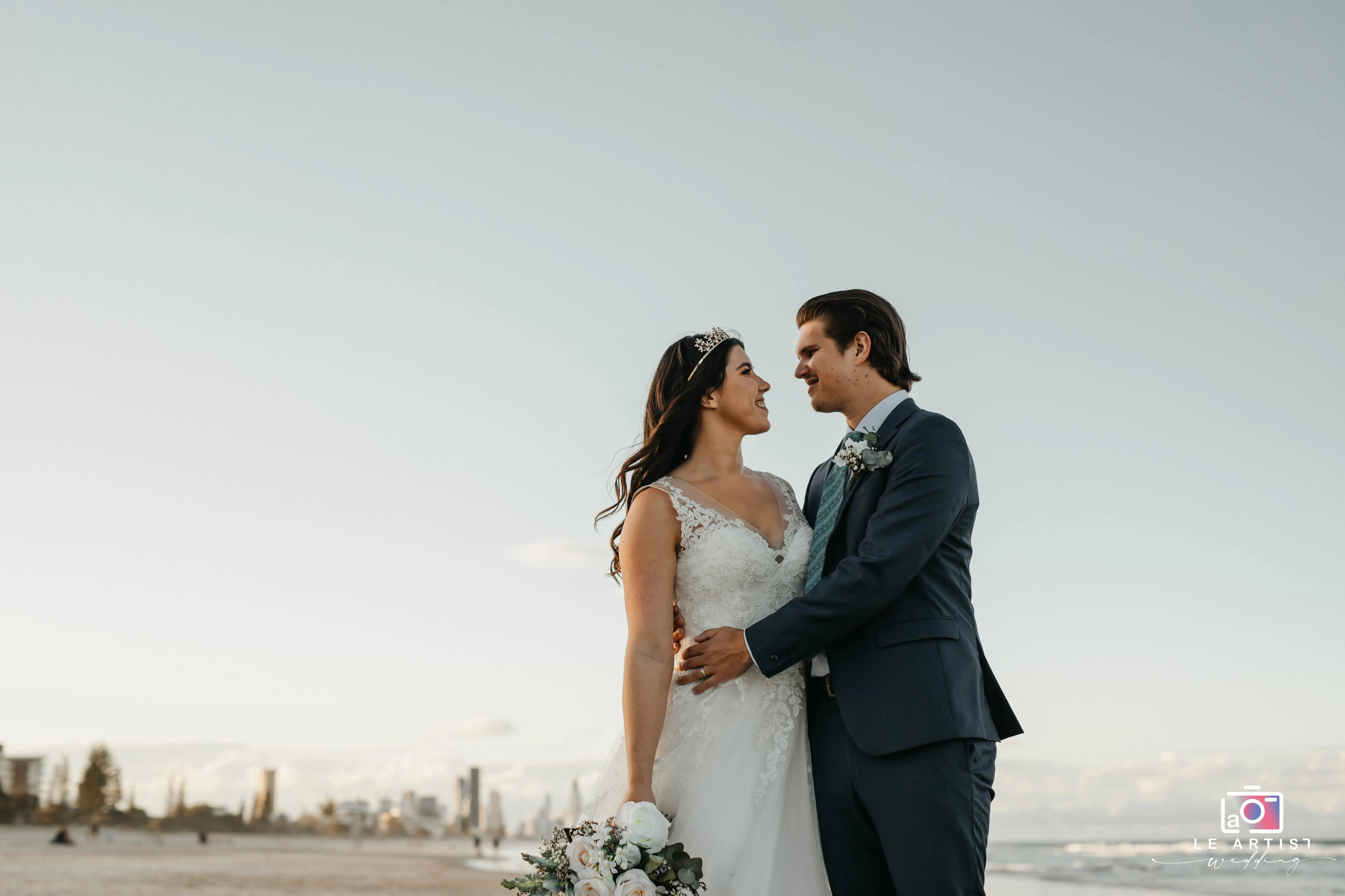 The newlywed couple shares an intimate moment on the beach, gazing lovingly into each other's eyes, with the city skyline in the background. Le Artist Wedding