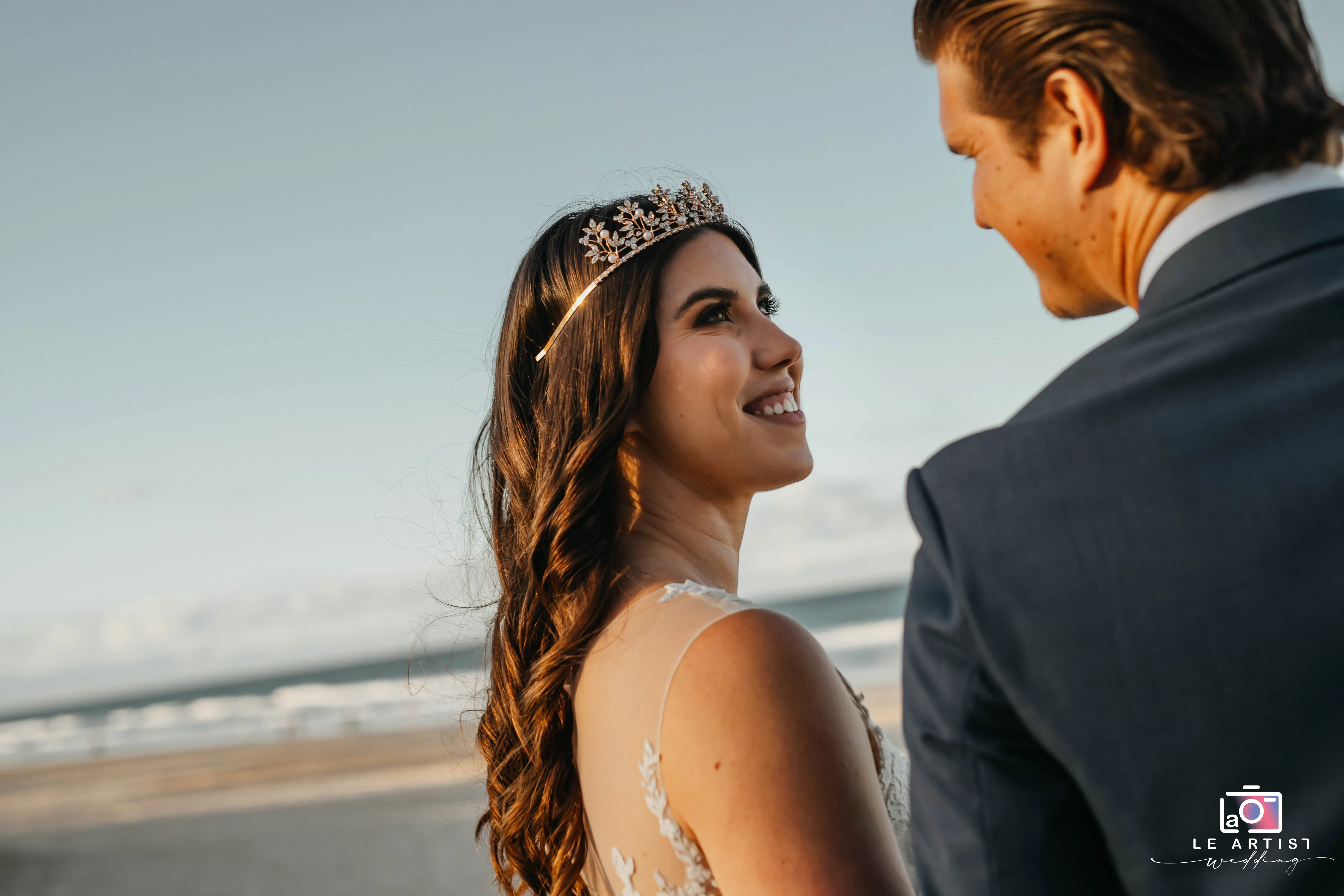 A joyful bride looks lovingly at her partner with the ocean in the background, capturing the essence of their special day. Le Artist Wedding