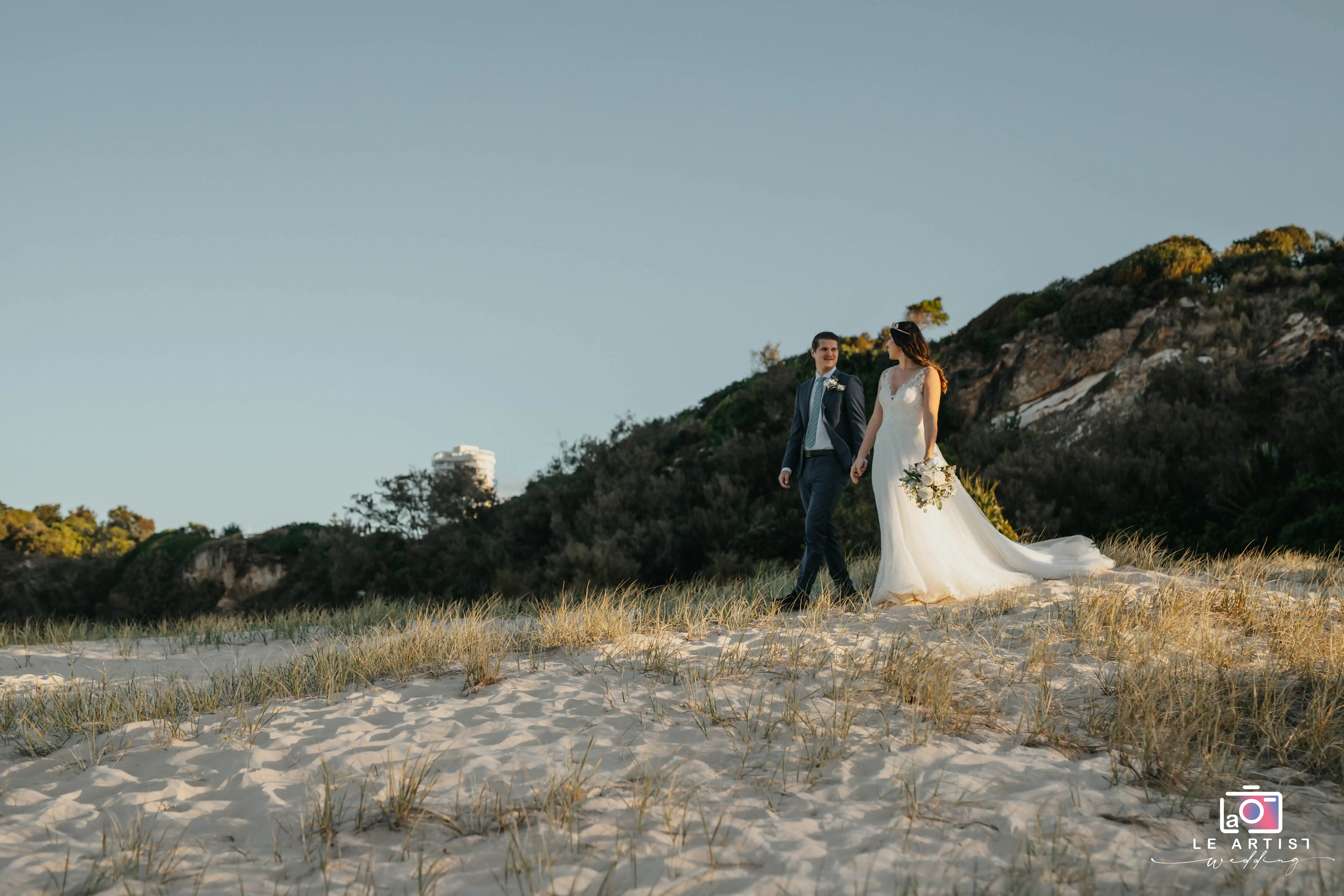 A newlywed couple strolls hand in hand along a scenic sandy beach, with the bride's train gently trailing behind and the early evening sunlight casting a warm glow. Le Artist Wedding