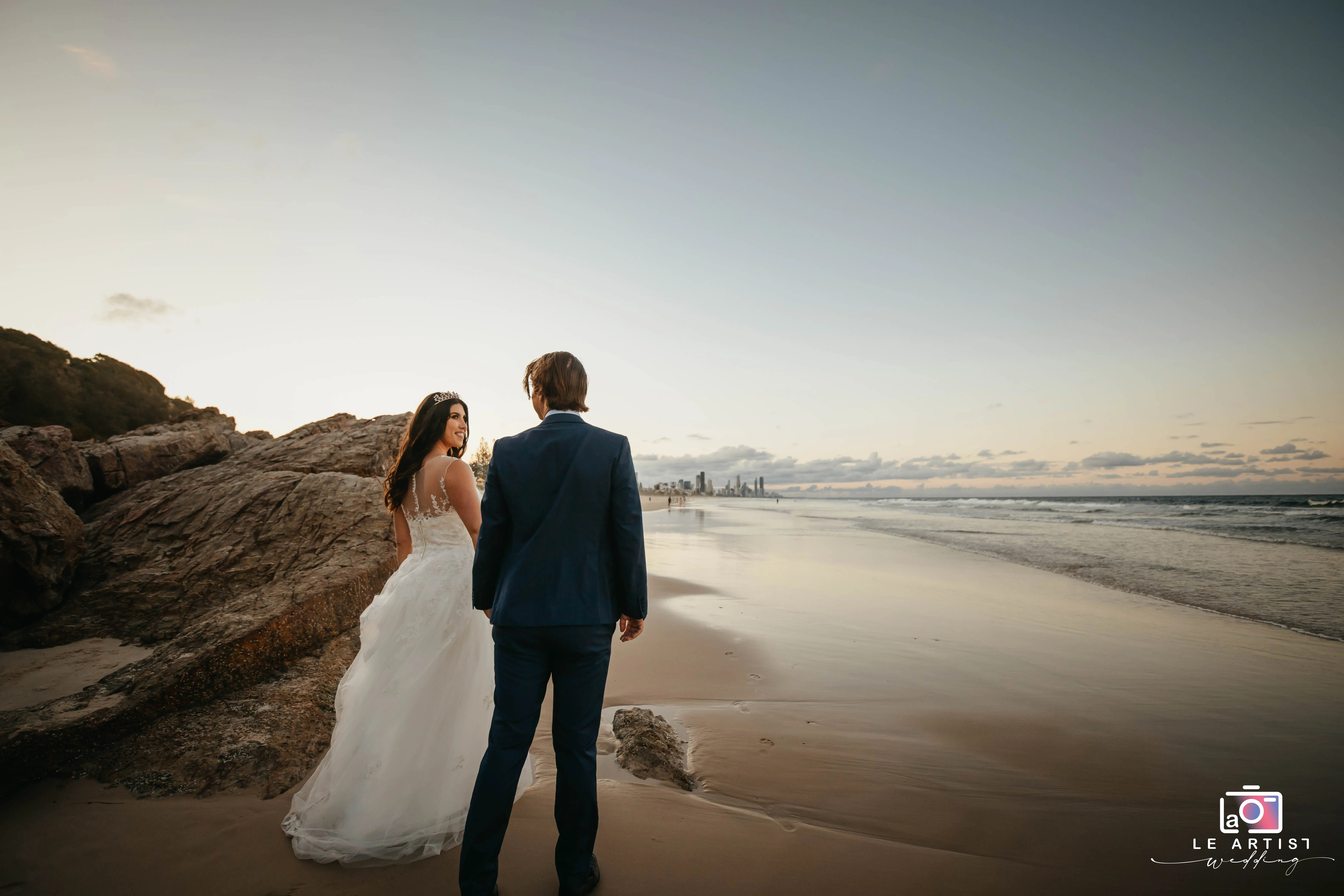 A newlywed couple strolls along a serene beach at sunset, capturing a perfect moment of joy and romance with the cityscape in the distance. Le Artist Wedding