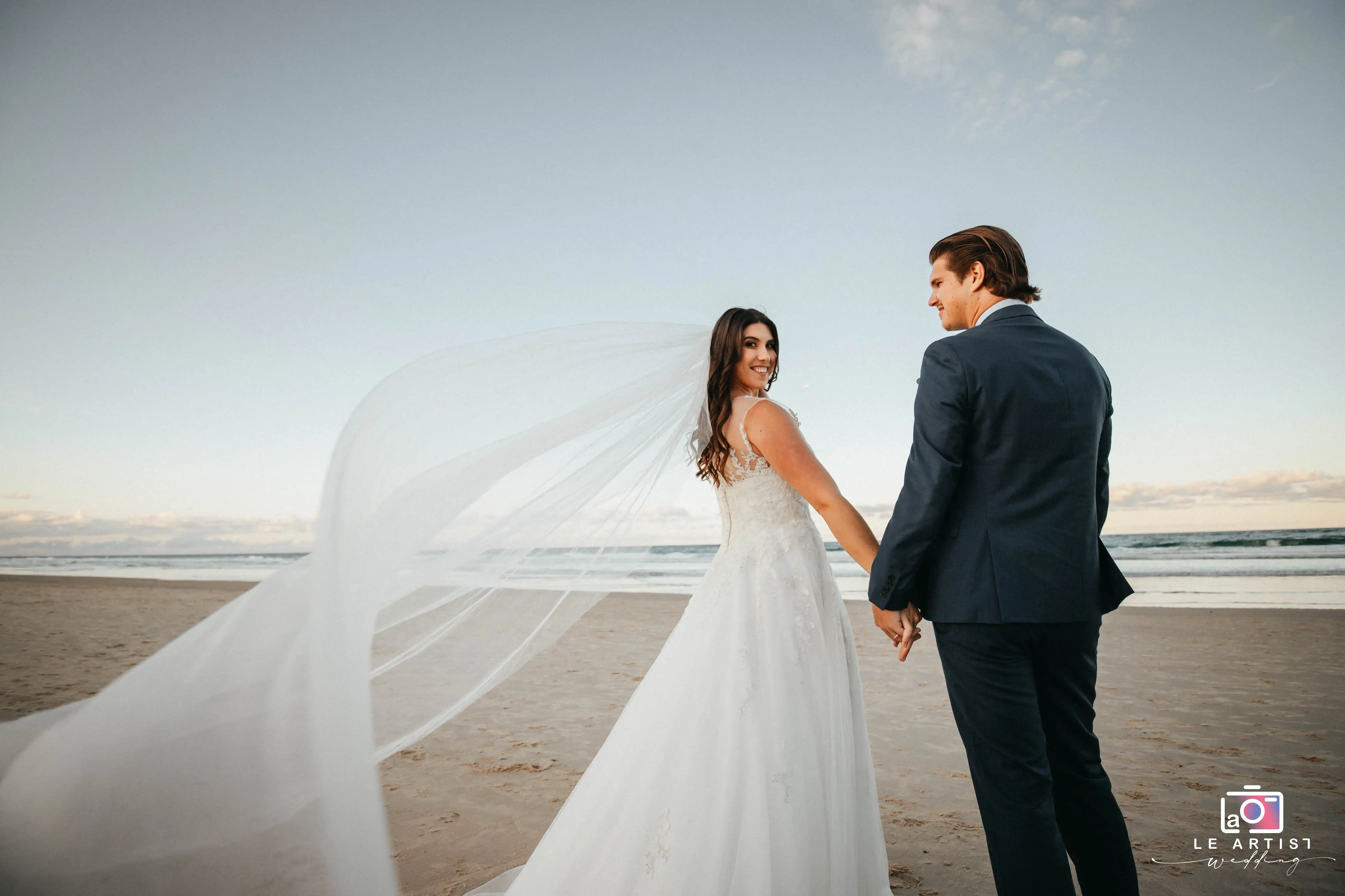A beautiful moment at the beach with the newlyweds holding hands, the bride's veil flowing in the breeze as they gaze at each other with smiles. Le Artist Wedding