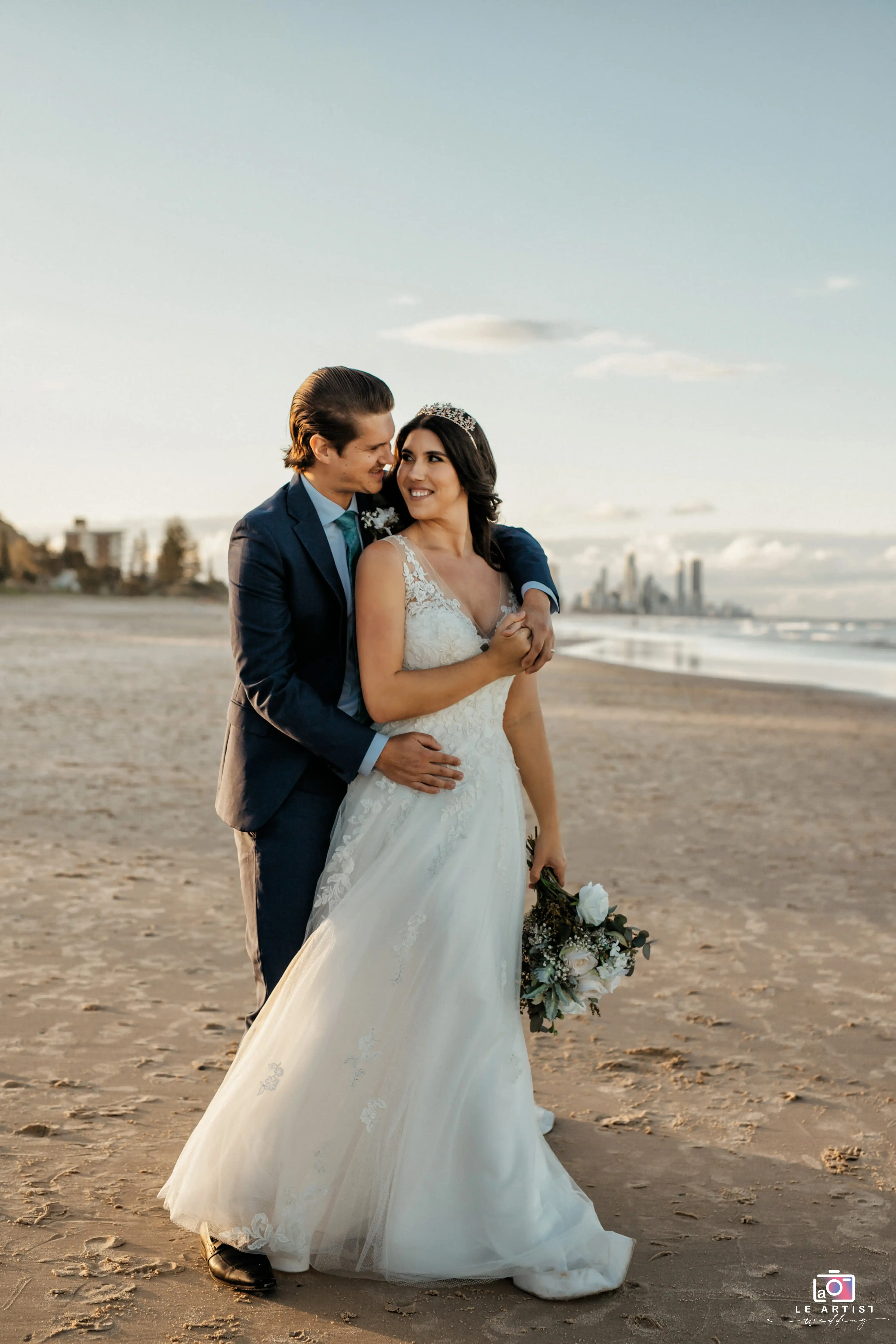 A romantic beachside moment of the newlyweds embracing, with the ocean and a distant cityscape as their backdrop. Le Artist Wedding