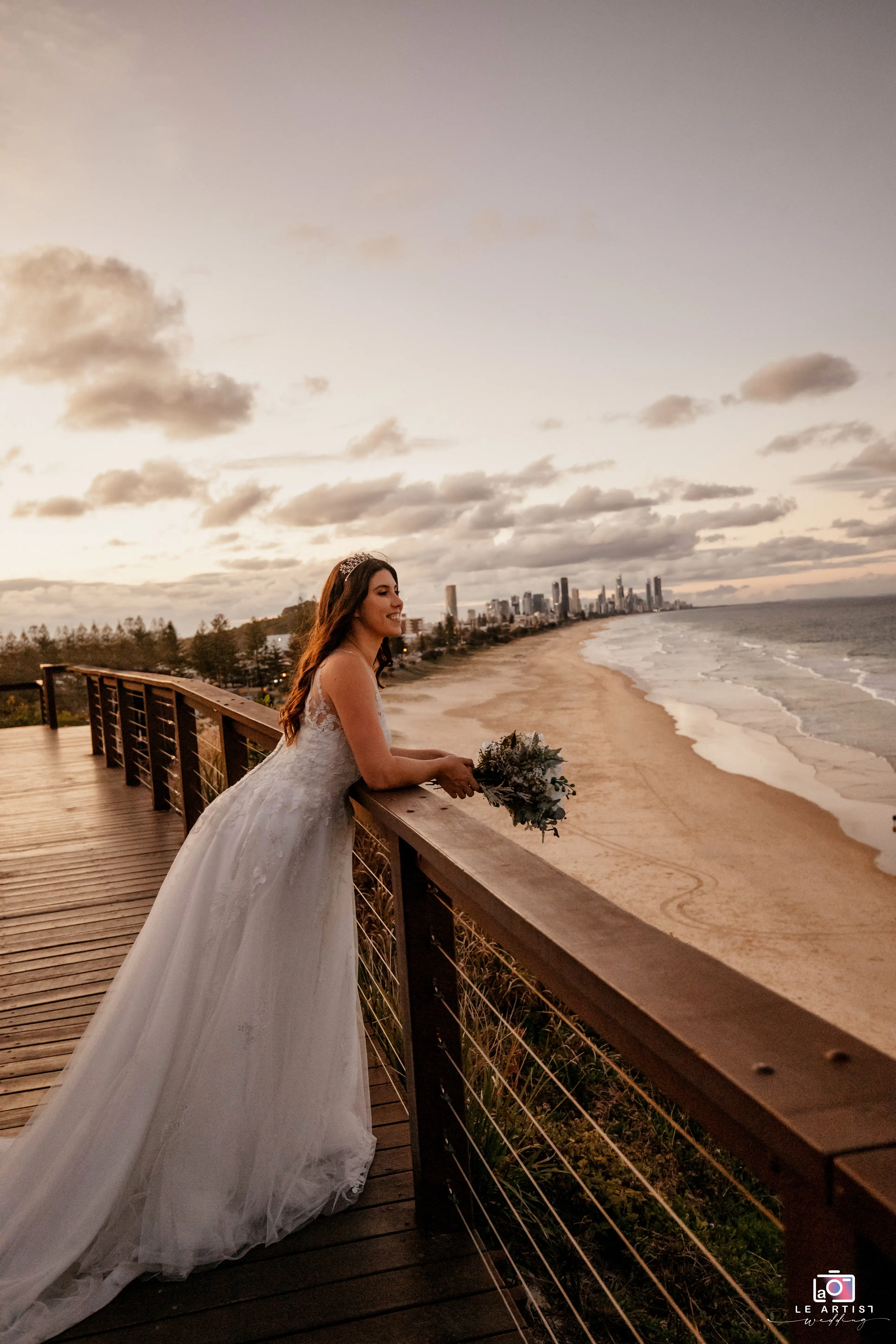 A bride gazes happily into the distance while holding her bouquet against a stunning beachside backdrop at sunset. Le Artist Wedding