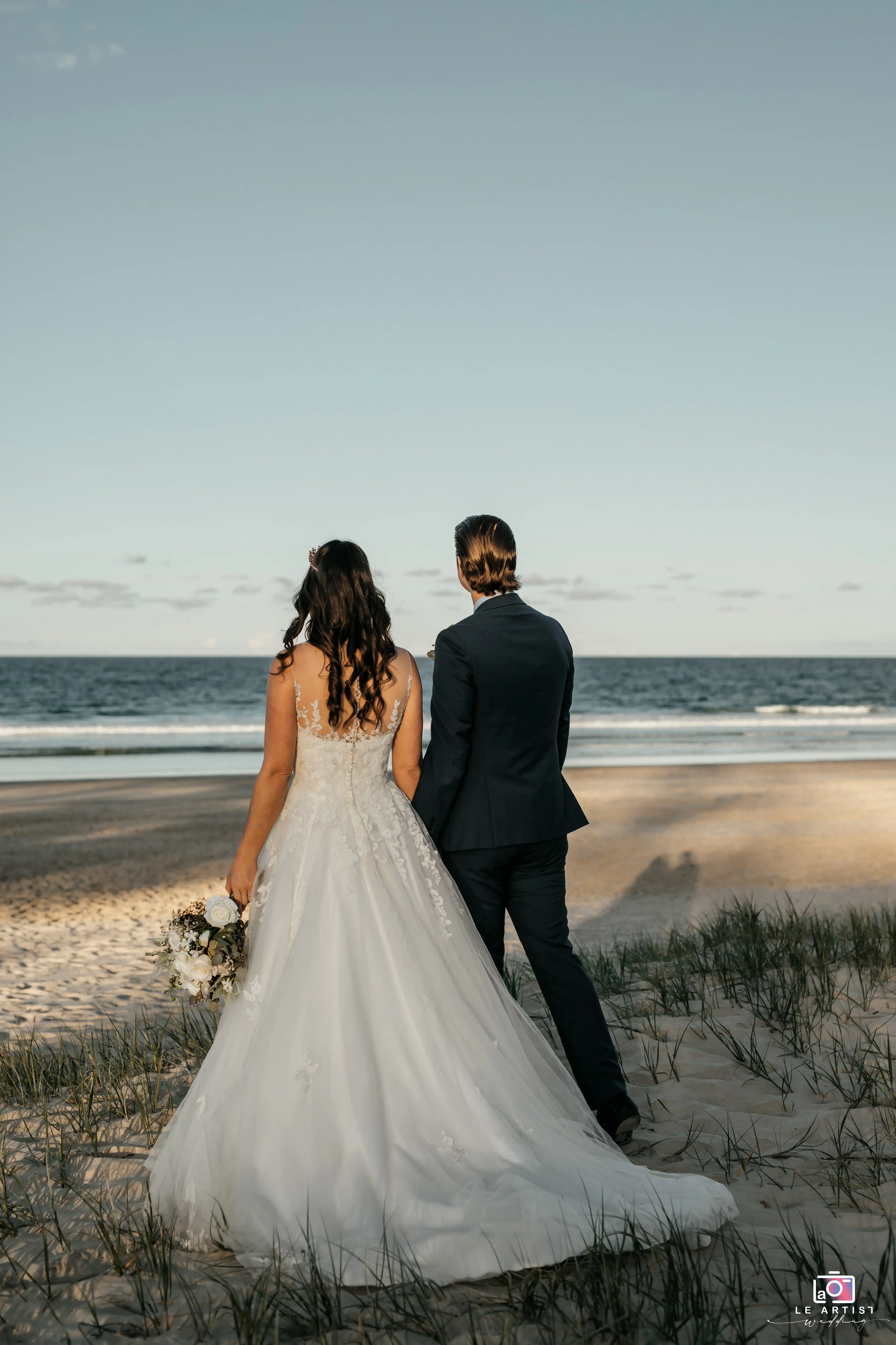 The bride and groom share a quiet moment together on the beach, gazing out towards the serene ocean. Le Artist Wedding