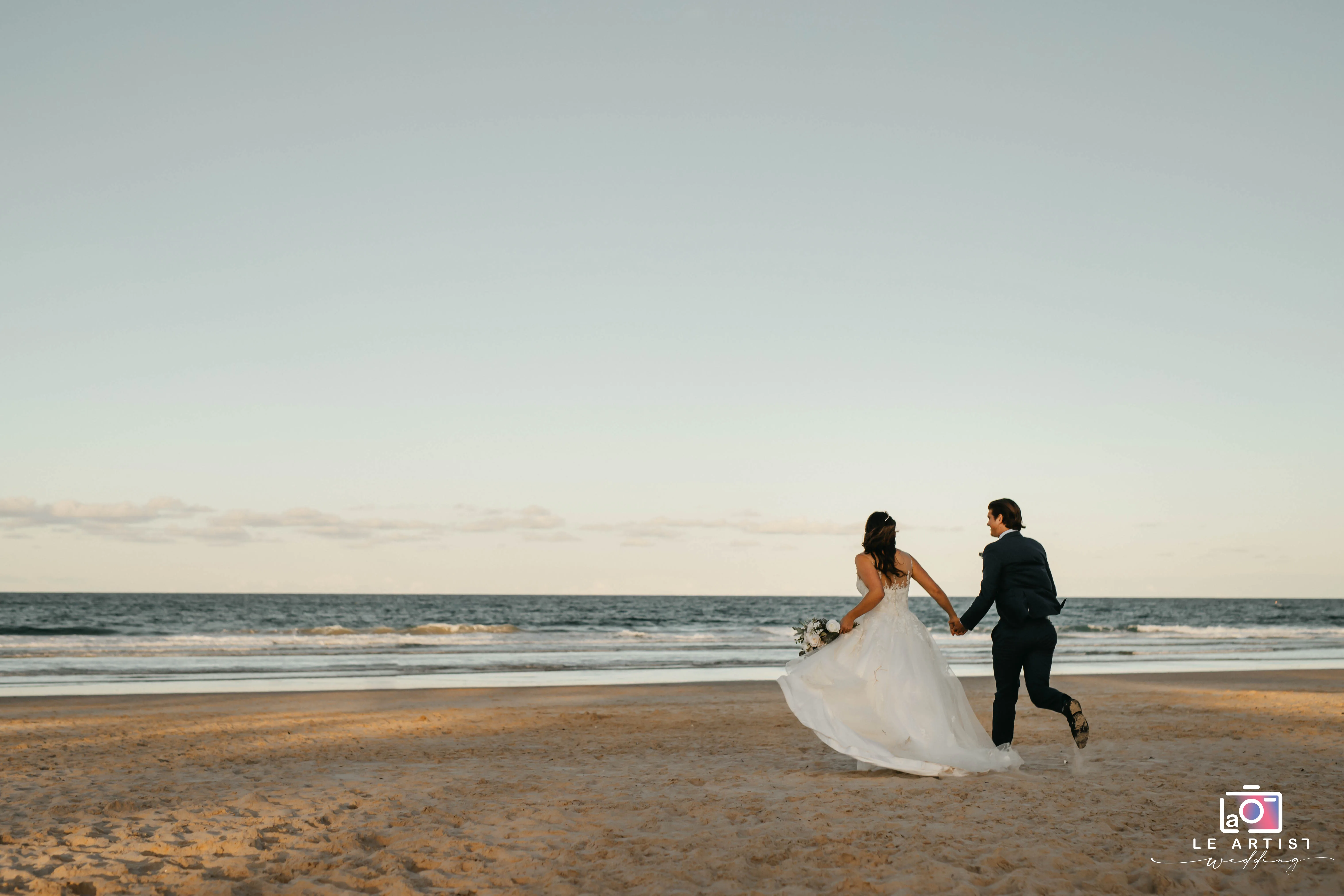 A bride and groom holding hands and running along the beach, capturing a joyful and carefree moment against the backdrop of the sea. Le Artist Wedding