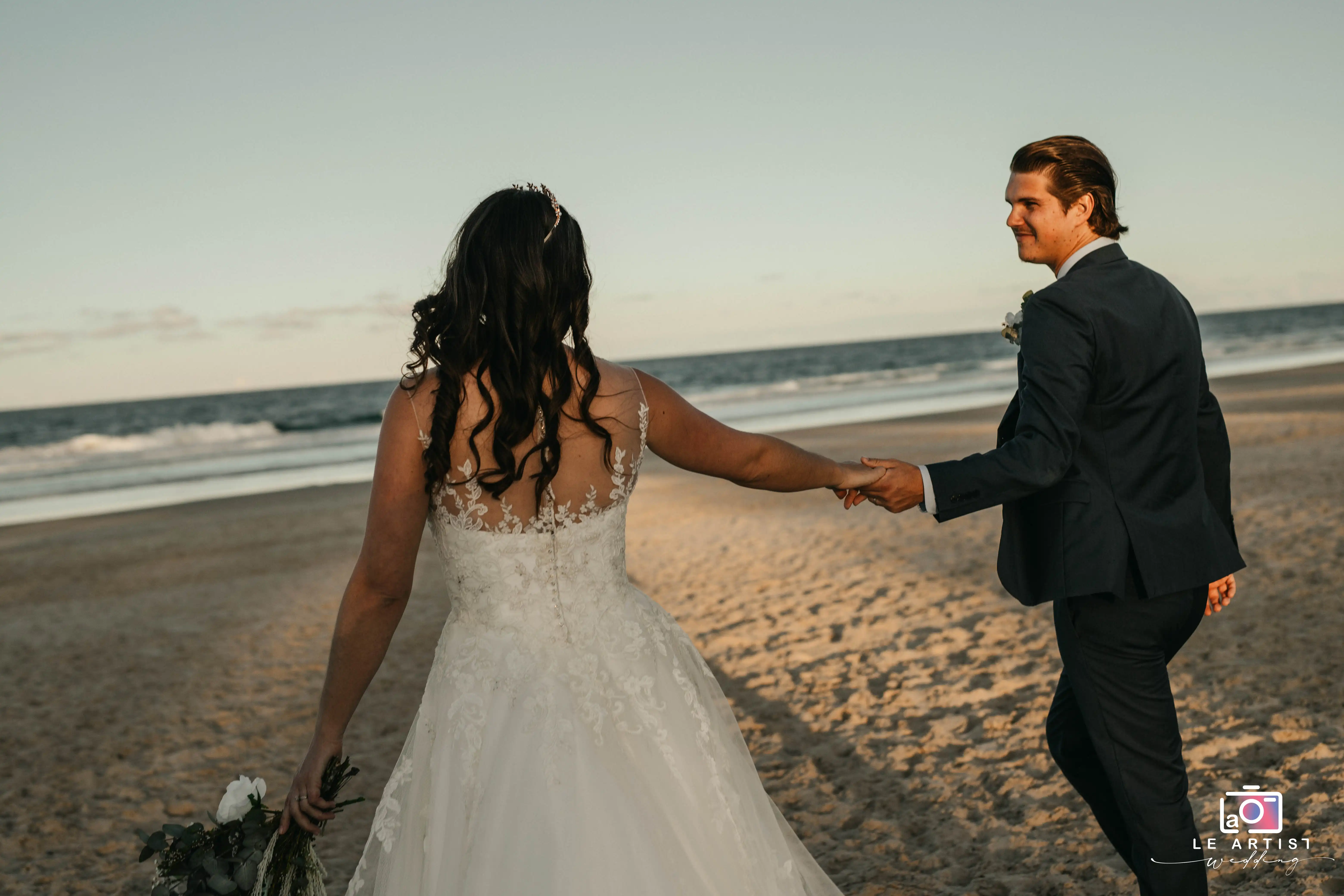A couple holds hands as they walk along the beach, embracing the beauty of their special day. Le Artist Wedding