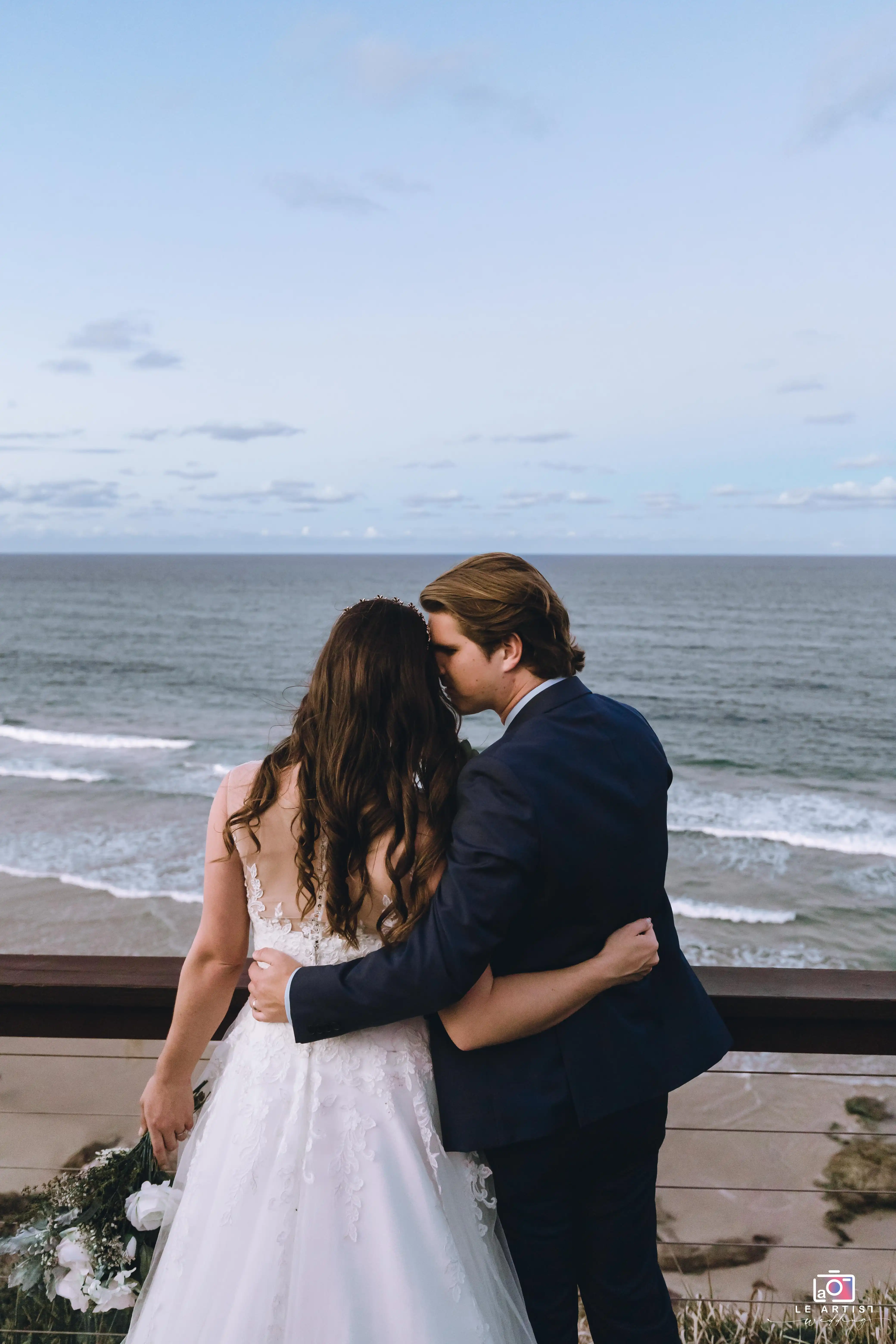 A couple shares a tender moment overlooking the ocean, capturing the essence of their special day. Le Artist Wedding