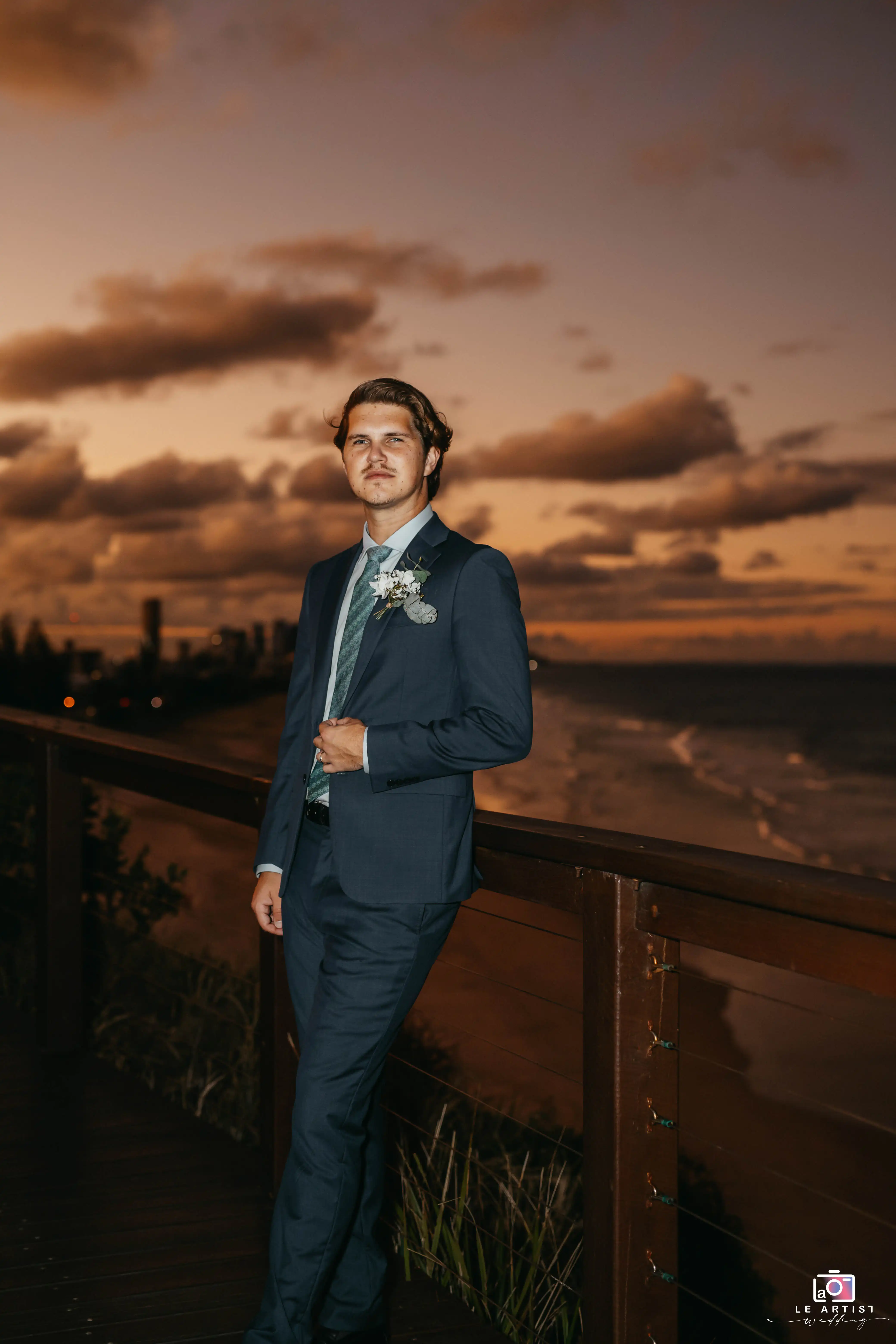 A groom stands on a wooden deck overlooking a serene beach at sunset, dressed in a classic dark suit with a floral boutonniere. Le Artist Wedding