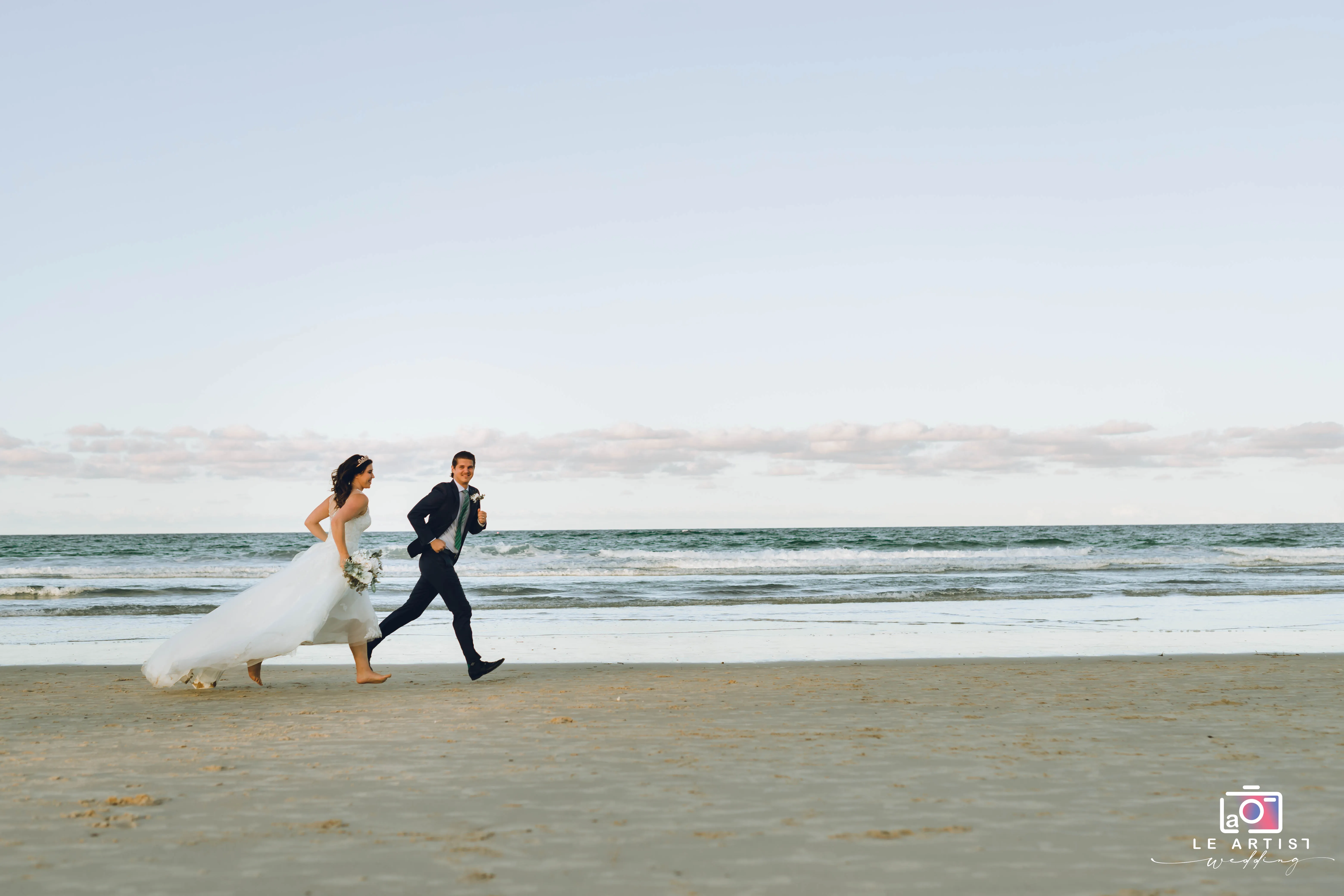 A joyous couple dashes barefoot along the serene shoreline, capturing a playful moment during their beach wedding. Le Artist Wedding
