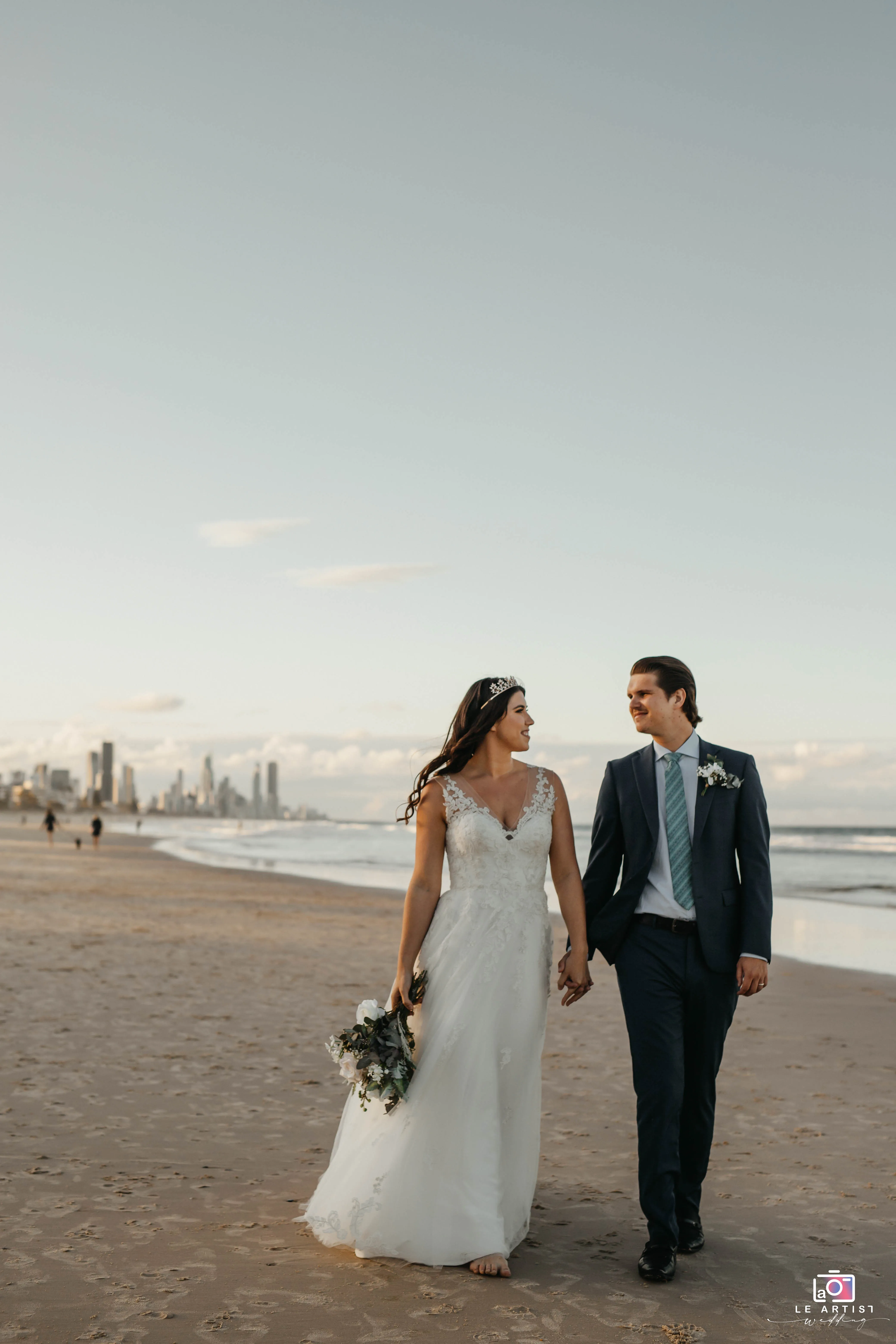 A newlywed couple walks hand-in-hand along a serene beach, sharing a moment of joy as the city skyline looms in the background. Le Artist Wedding