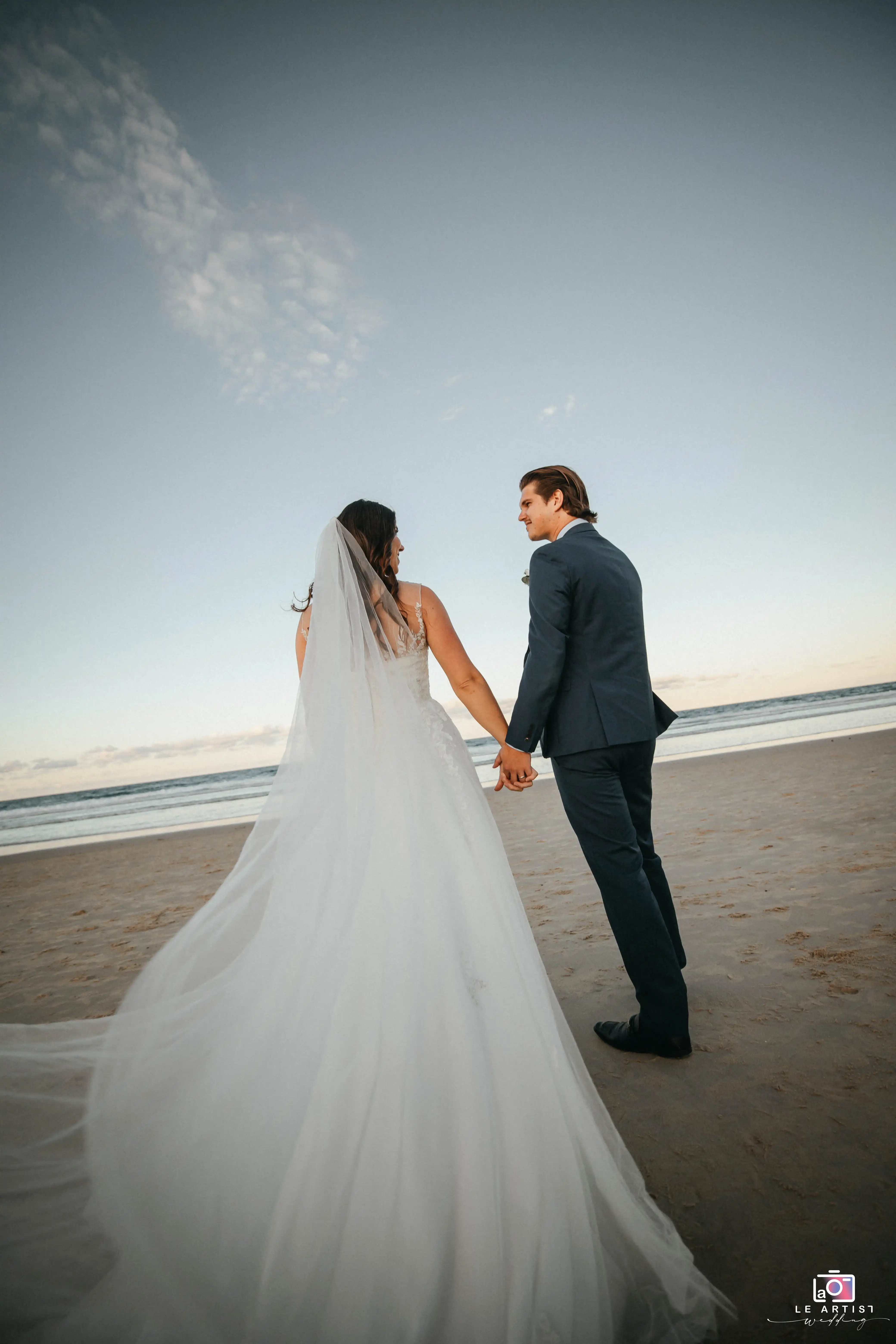 The newlyweds share a romantic moment, hand in hand, as they walk along the serene beach, their expressions full of love and joy. Le Artist Wedding