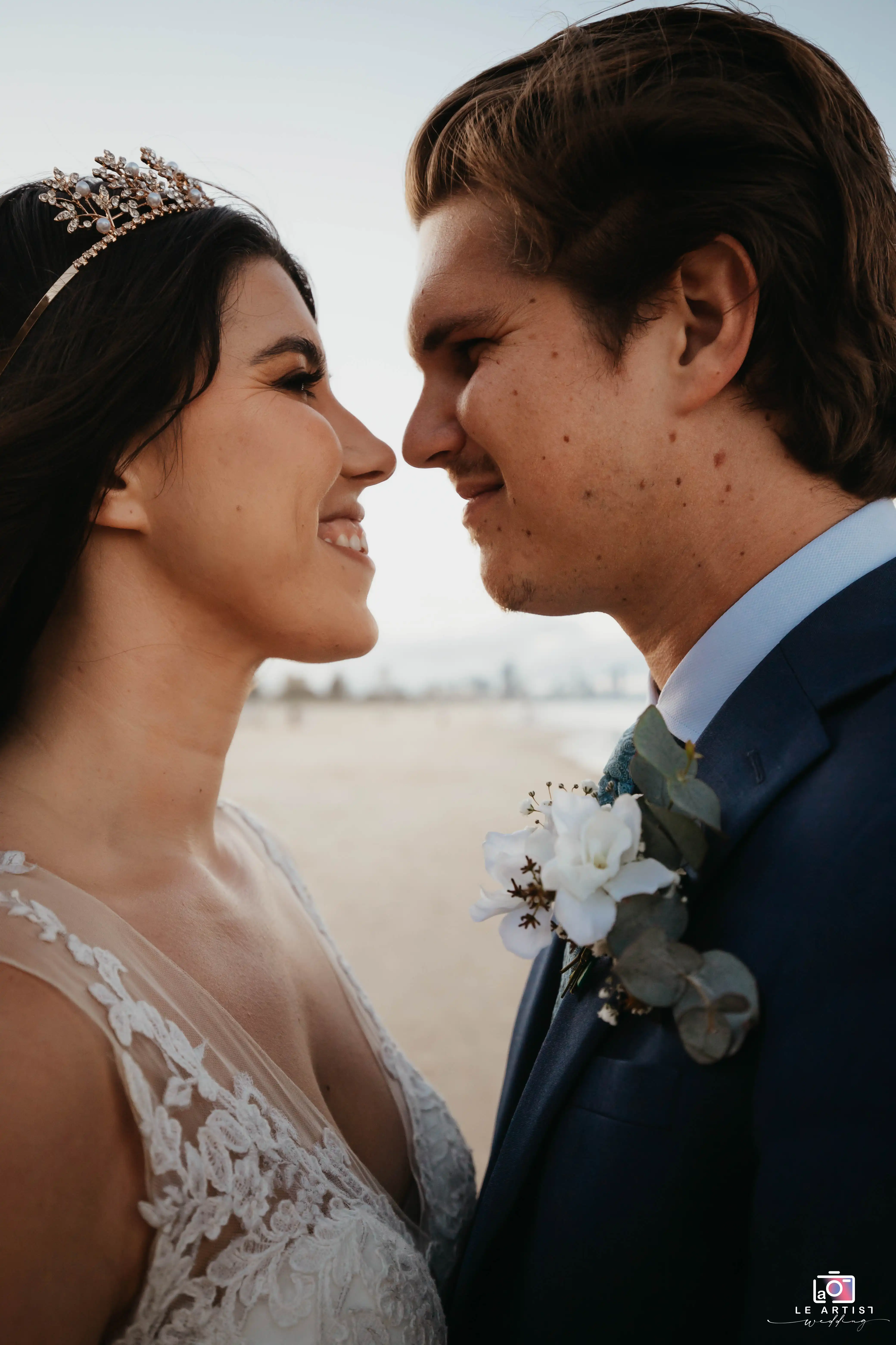 A romantic close-up of the couple sharing a tender moment on the beach, with the bride wearing a delicate tiara and the groom in a sharp blue suit adorned with a floral boutonniere. Le Artist Wedding