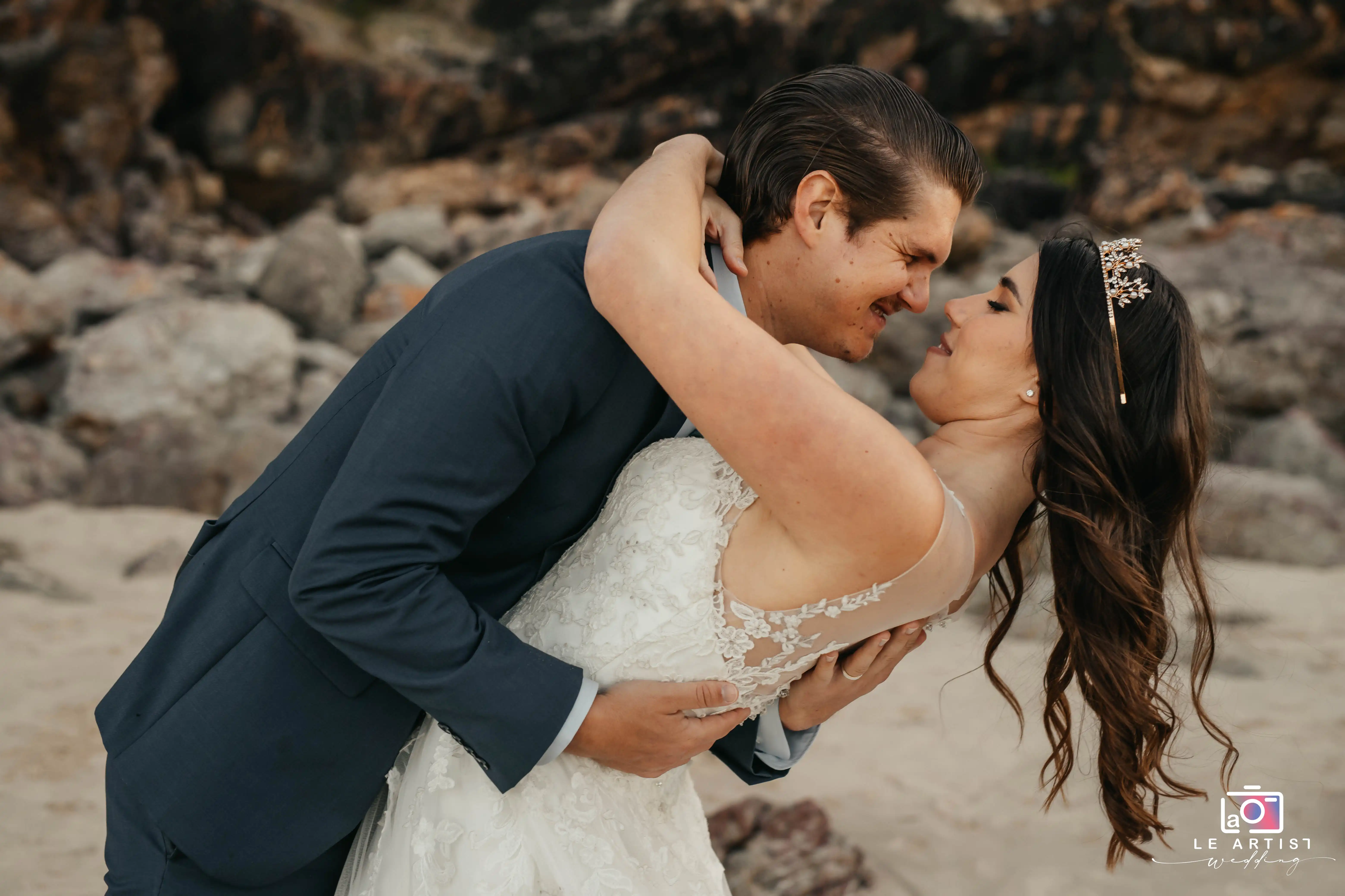 A romantic moment captured as the couple shares a joyous embrace on a rocky beach setting, highlighting their happiness and love. Le Artist Wedding