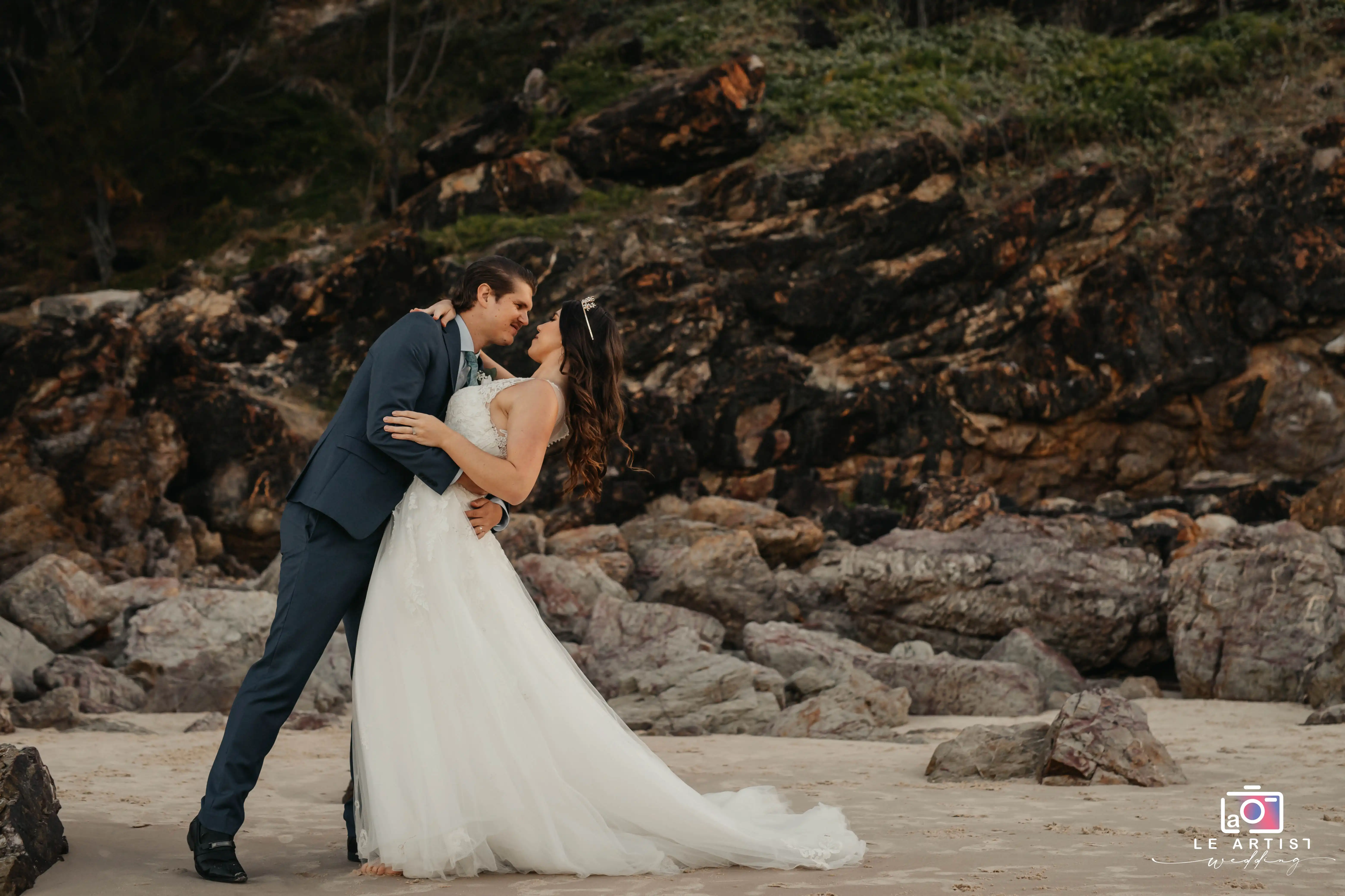 A romantic moment captured as the bride and groom share an intimate embrace on a rocky beach. Le Artist Wedding