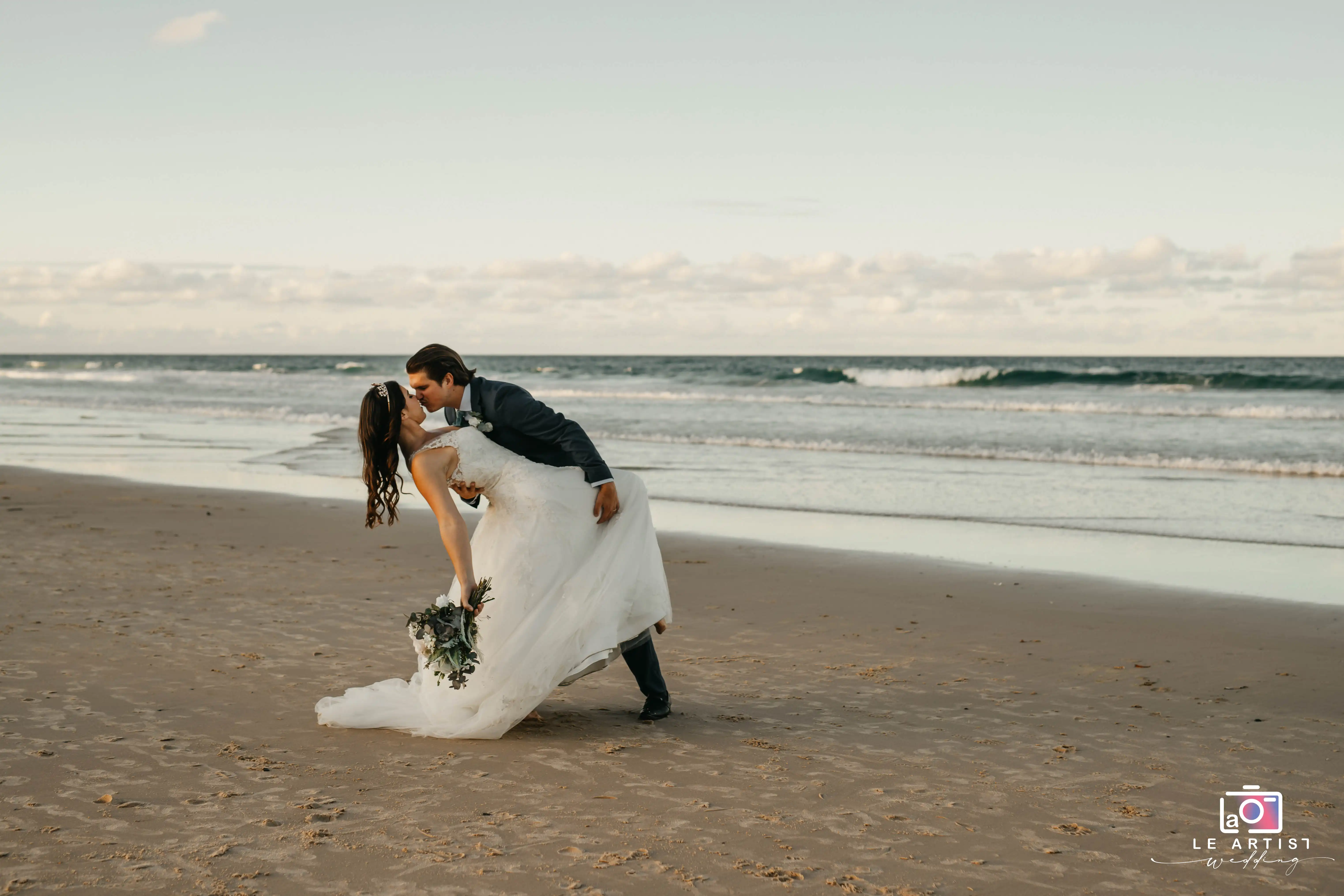 A romantic beachside kiss captured during a wedding photo session, with the couple elegantly posed against the backdrop of gentle waves and an open sky. Le Artist Wedding