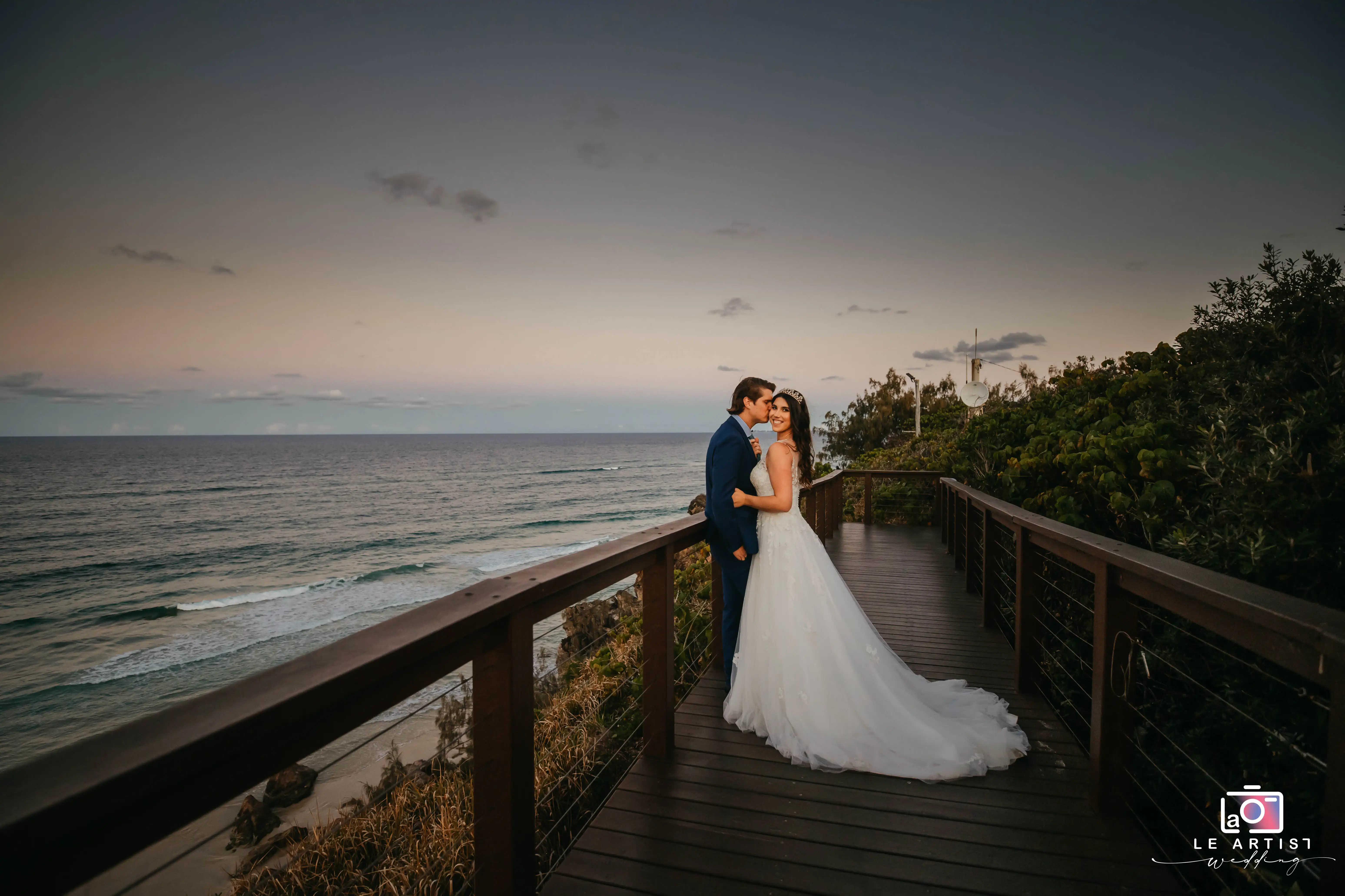 A couple shares a romantic moment on a scenic boardwalk overlooking the ocean at sunset, highlighting the serene beauty of their wedding day. Le Artist Wedding