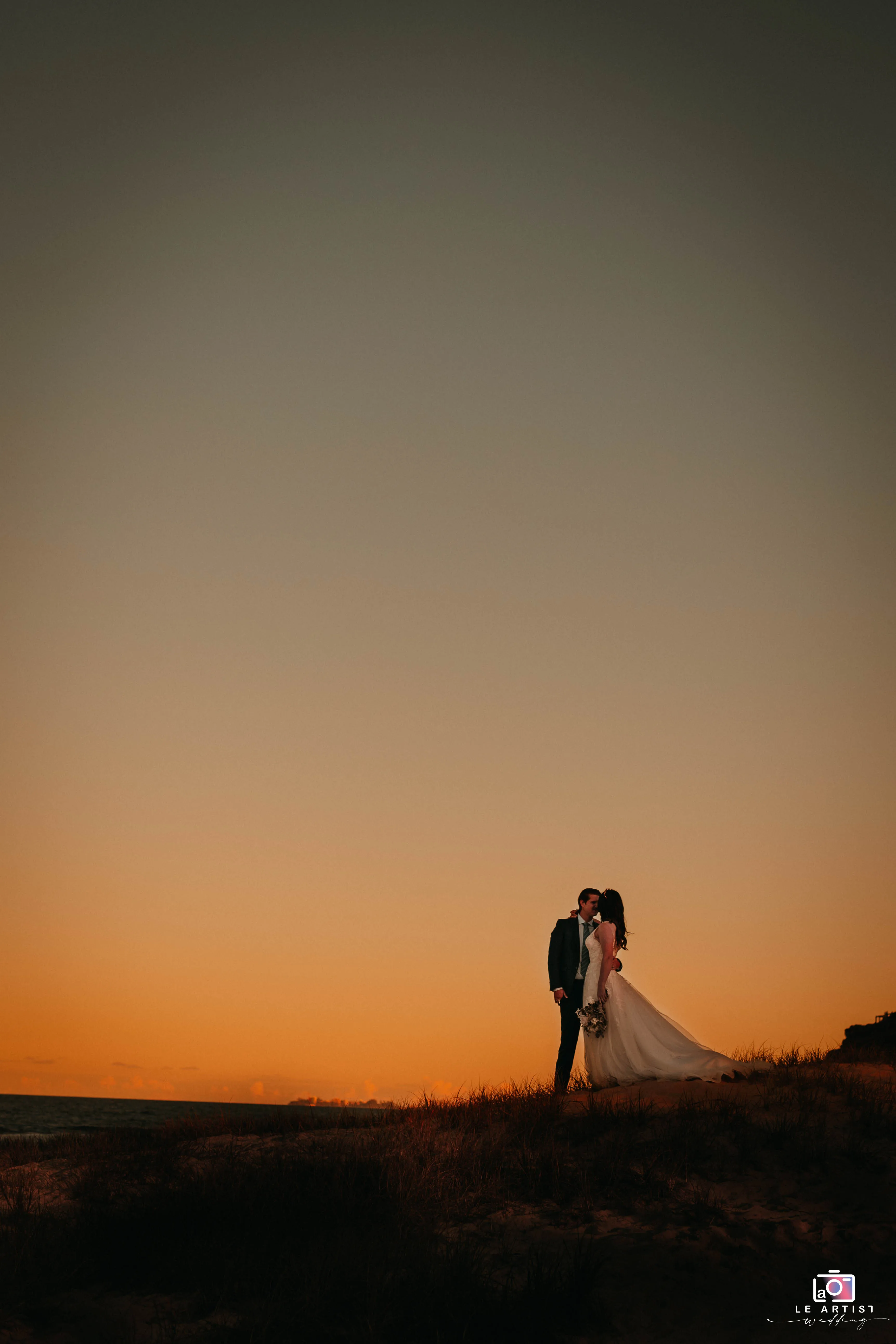 A couple shares a romantic moment on a hillside with a stunning sunset backdrop, capturing the essence of their special day. Le Artist Wedding