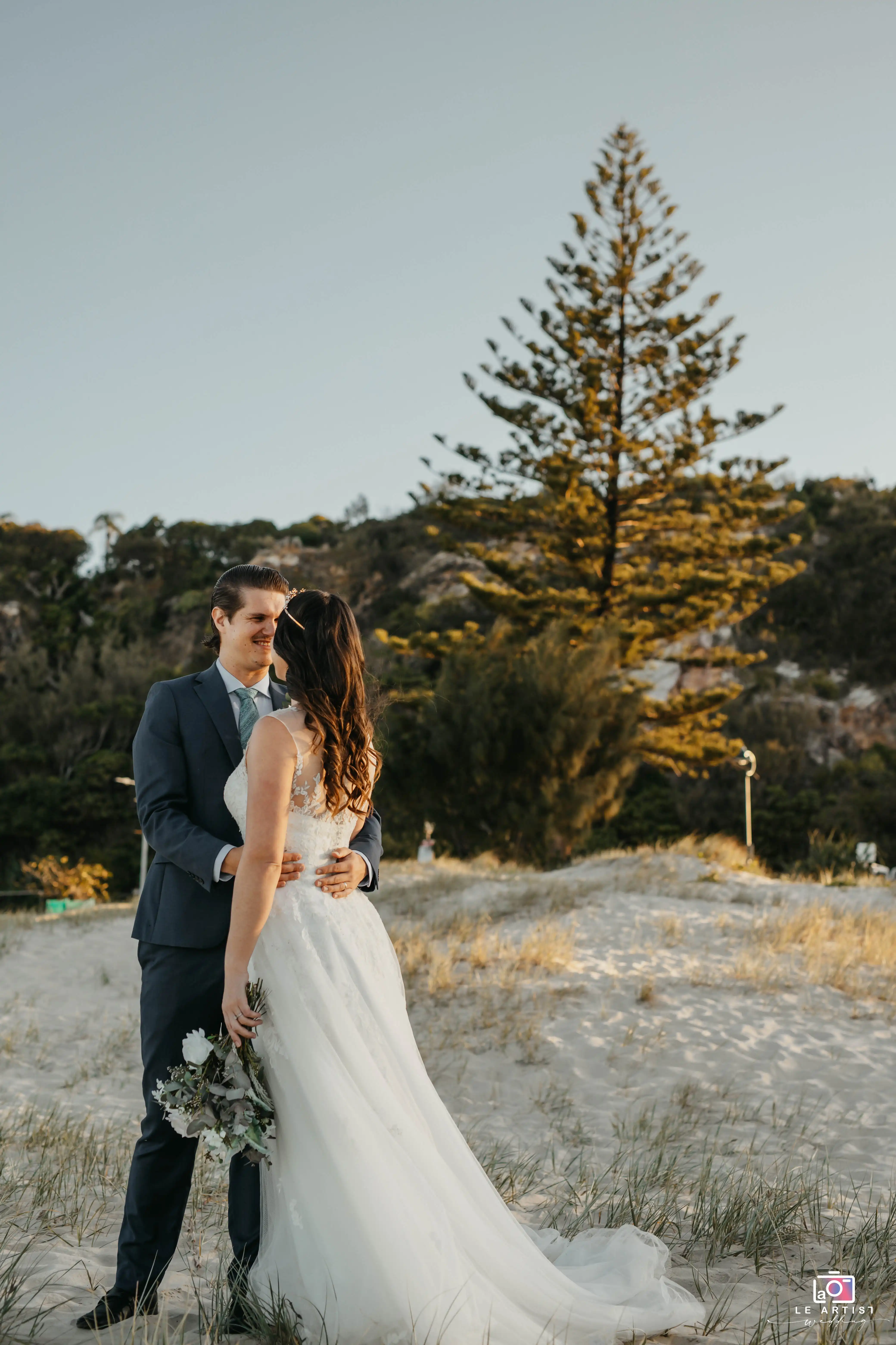 The couple shares a tender moment on a sunlit sandy landscape, surrounded by nature's tranquility. Le Artist Wedding