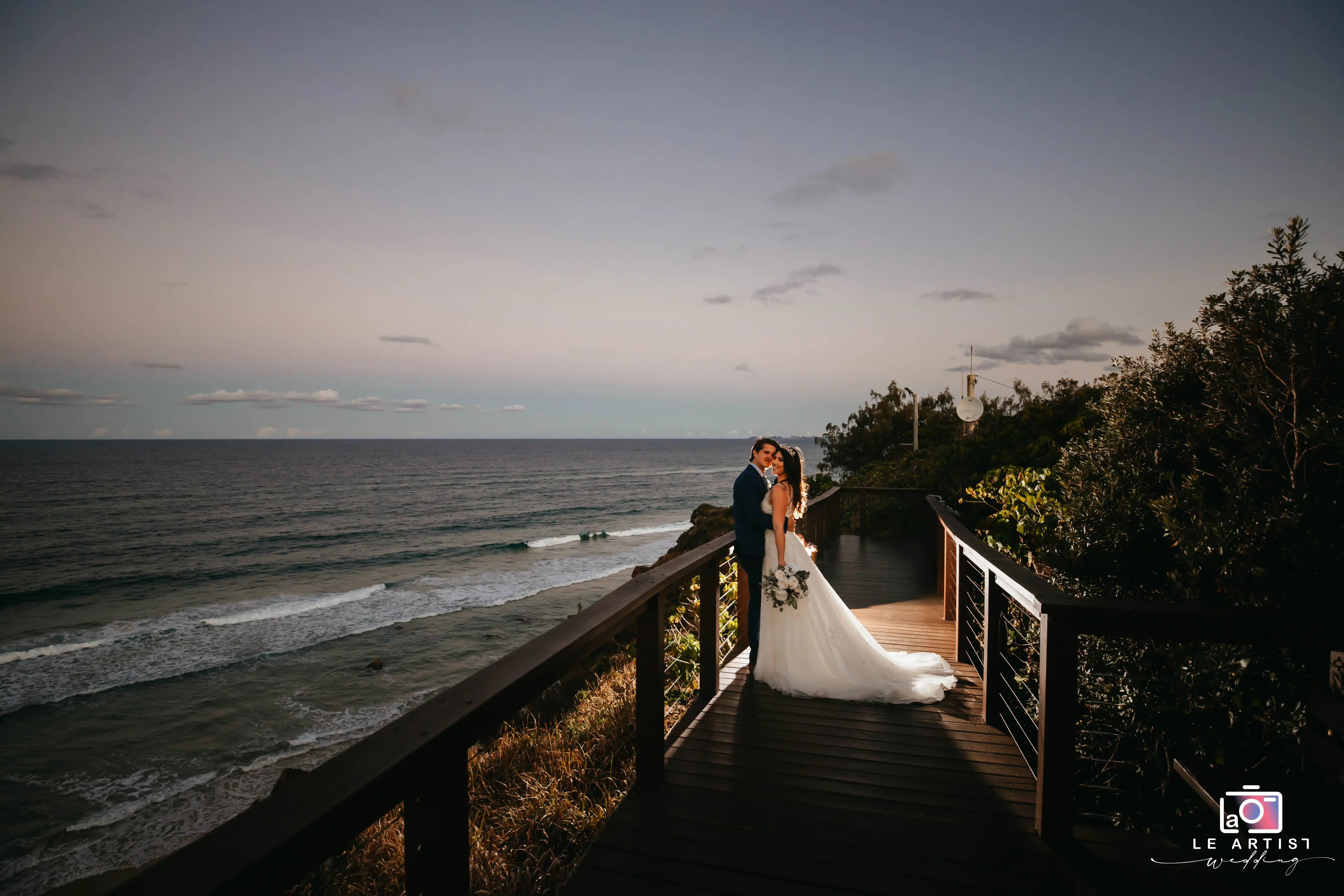 A romantic sunset moment as the newlyweds share a kiss on a coastal boardwalk, with the ocean waves softly breaking in the background. Le Artist Wedding