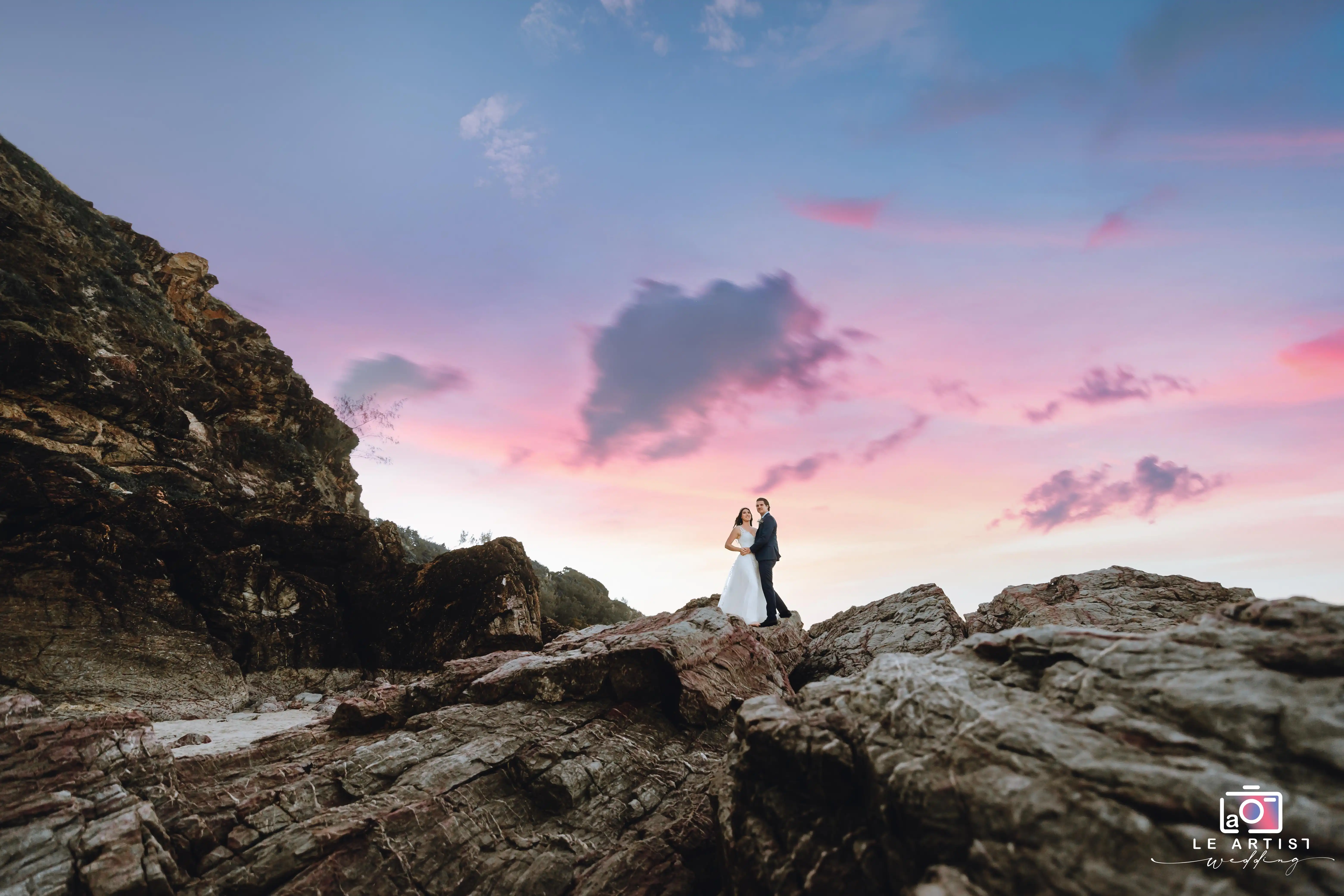 A couple embraces on rocky terrain against a stunning sunset sky, capturing a moment of romantic serenity and natural beauty. Le Artist Wedding